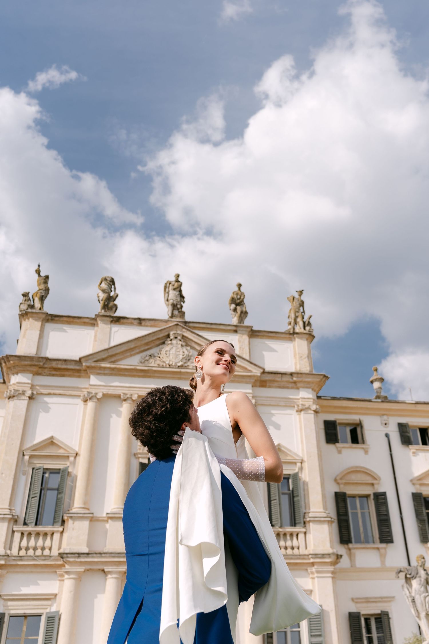 A couple in wedding attire shares a romantic moment in front of an ornate white building with classical architecture. Villa Mosconi Bertani