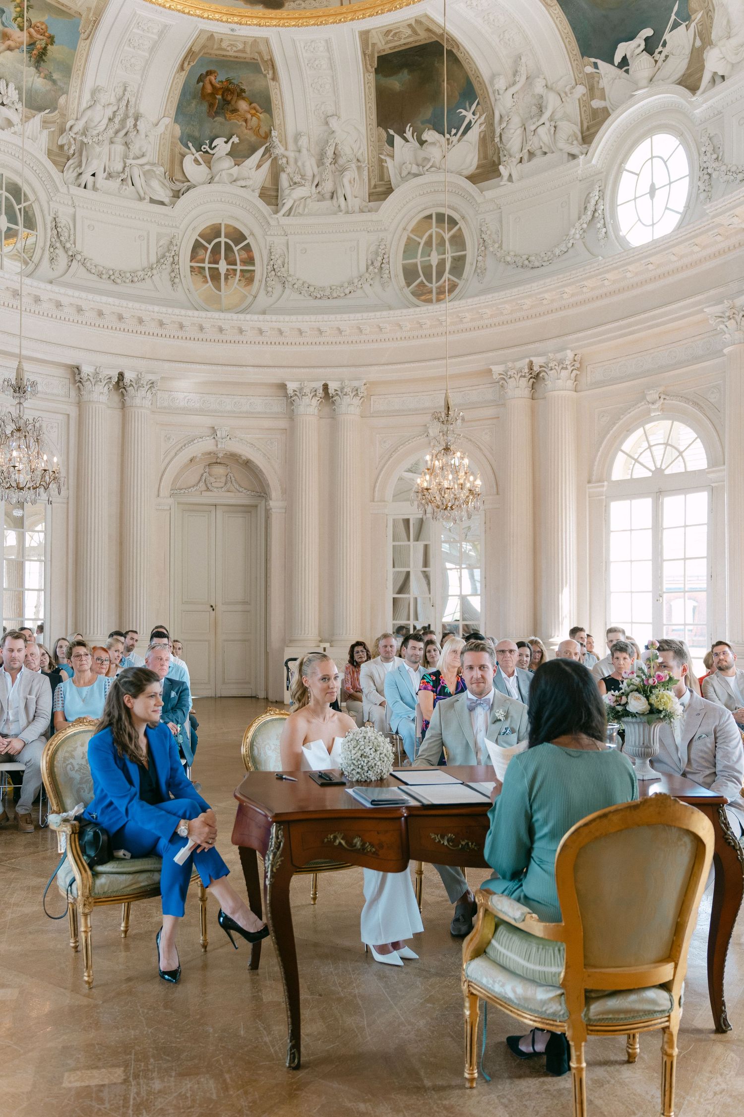Elegant ceremony taking place in an ornate white baroque room with crystal chandeliers and arched windows. Schloss Solitude