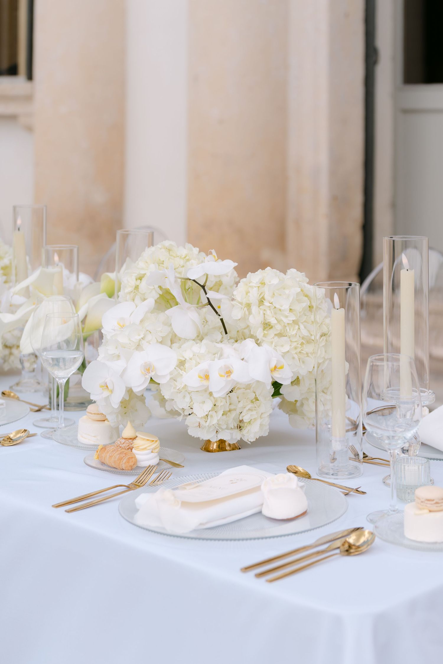 Elegant white wedding table setting with hydrangeas, candles, and gold flatware on crisp white linens. Villa Mosconi Bertani