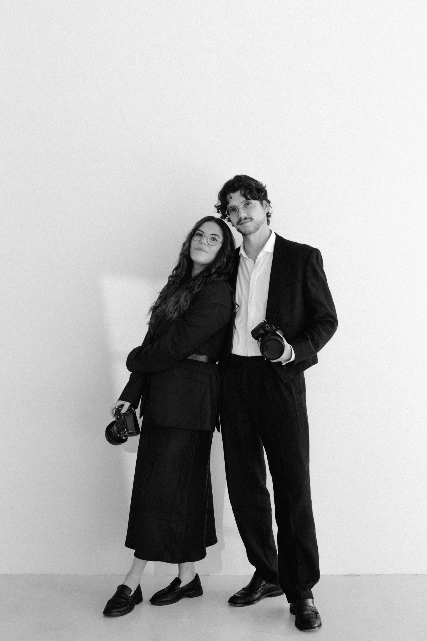 Black and white photo of a stylish couple in suits holding cameras, posing against a white wall.