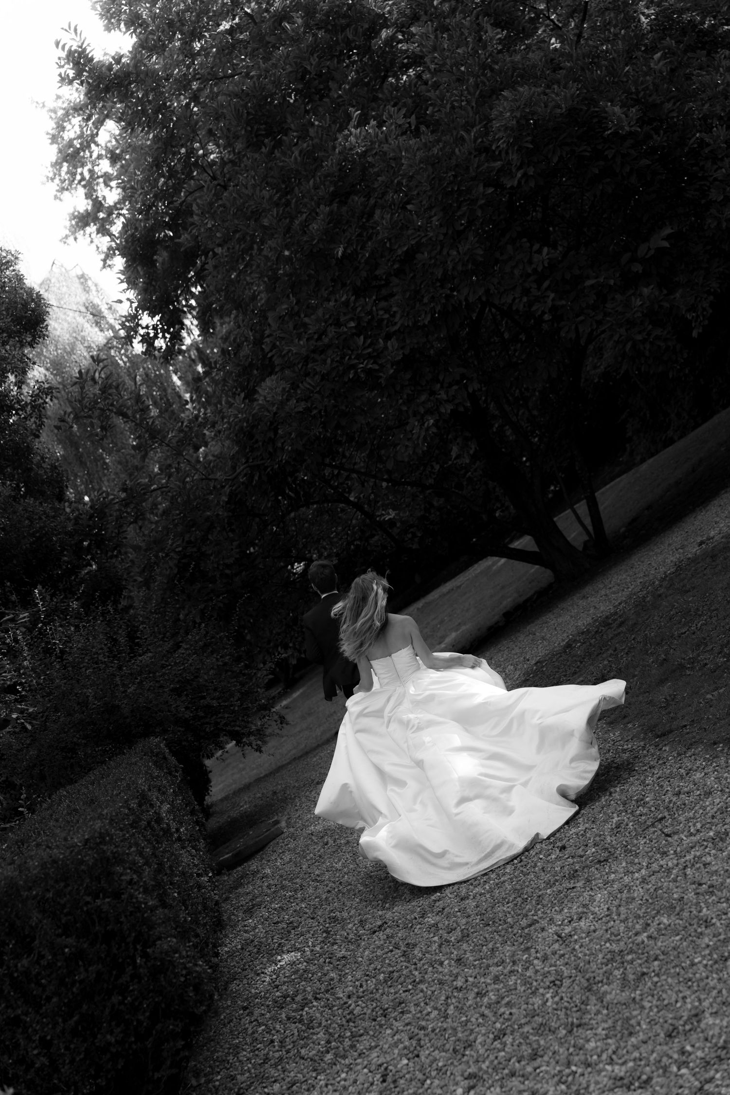 Black and white photo of a bride in a flowing white wedding gown running across a grassy hillside beside dark trees.