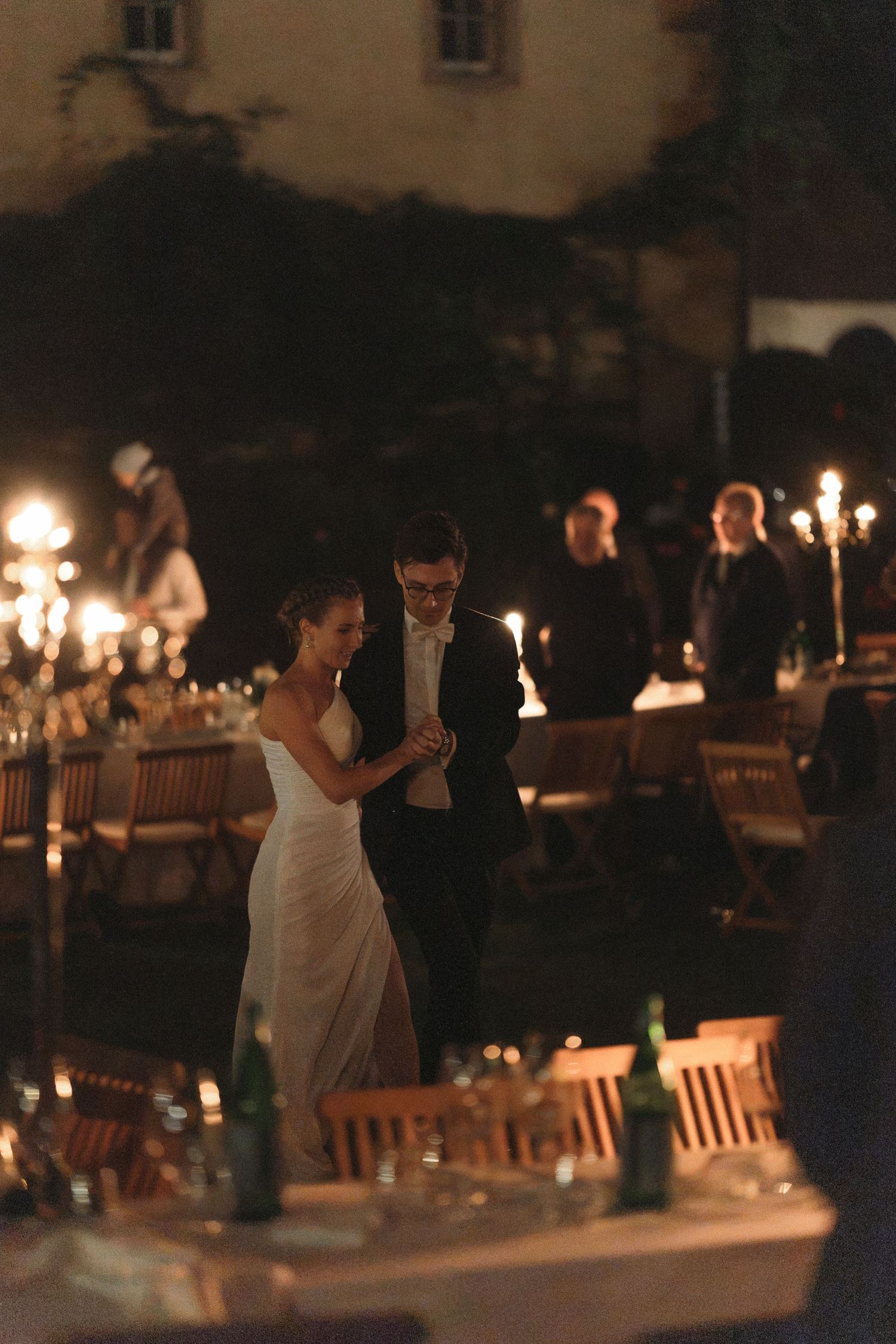 Bride in a white gown and groom in a dark suit dance outdoors at an evening wedding reception lit by candelabras and surrounded by guests.