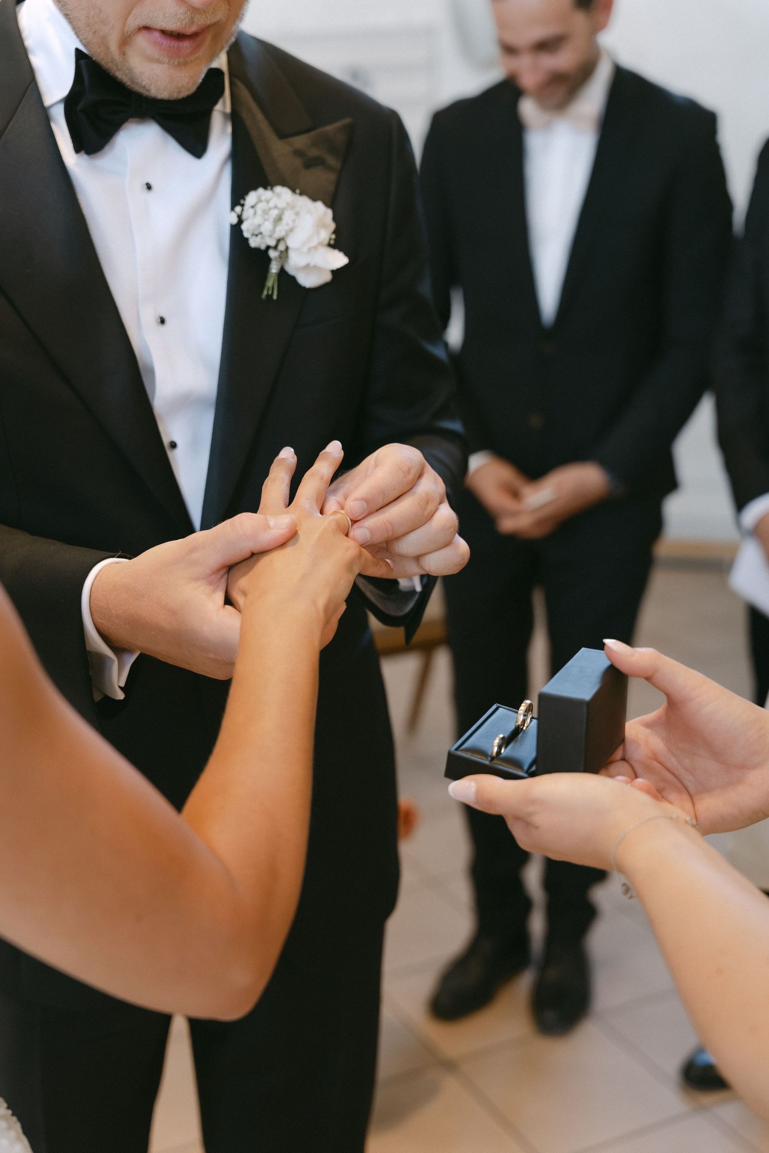 Groom in a black tuxedo and white boutonniere slides a wedding ring onto his bride's finger during the ceremony as guests look on.