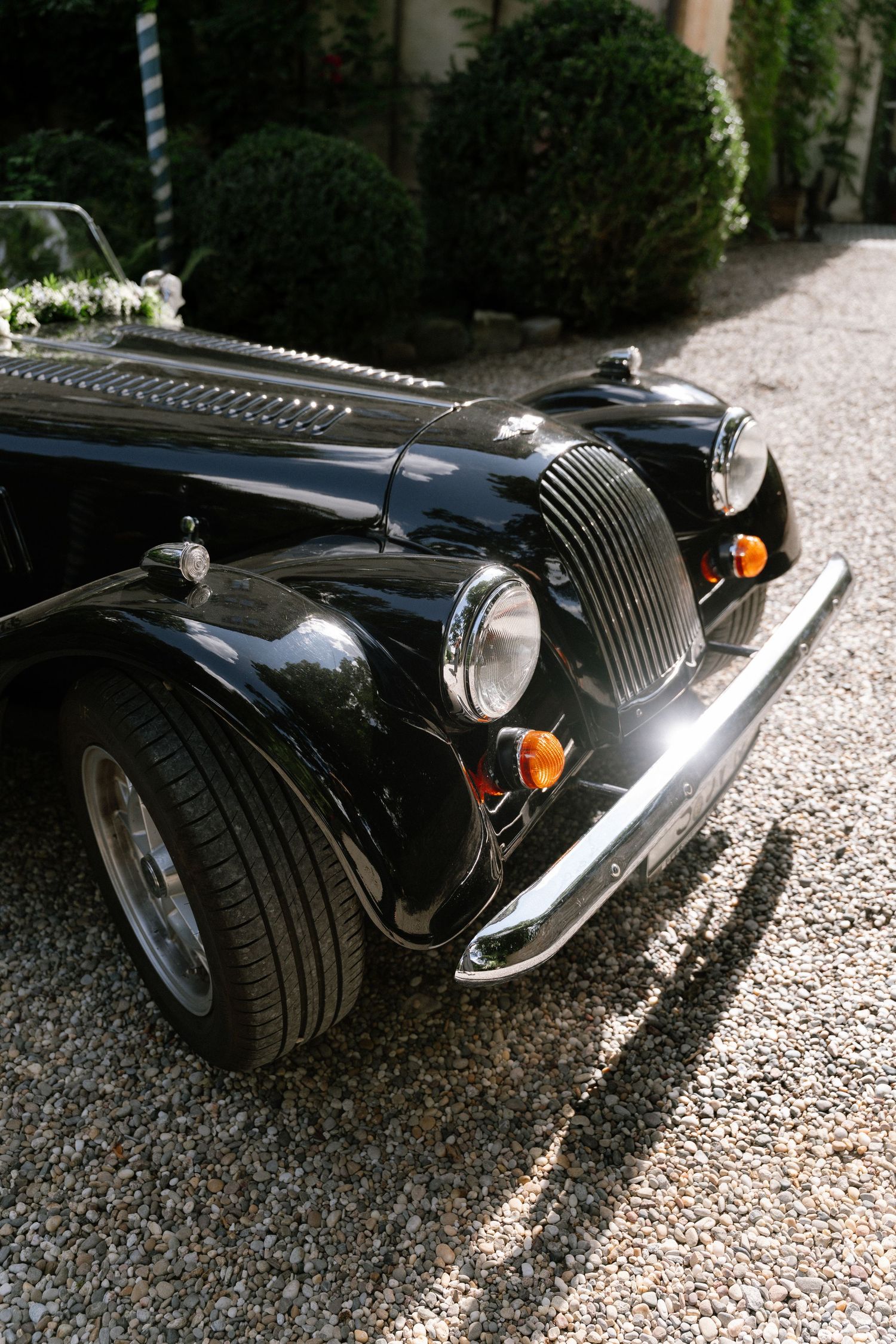 Black vintage Morgan roadster with chrome grille and round headlights parked on a gravel driveway beside trimmed green hedges.