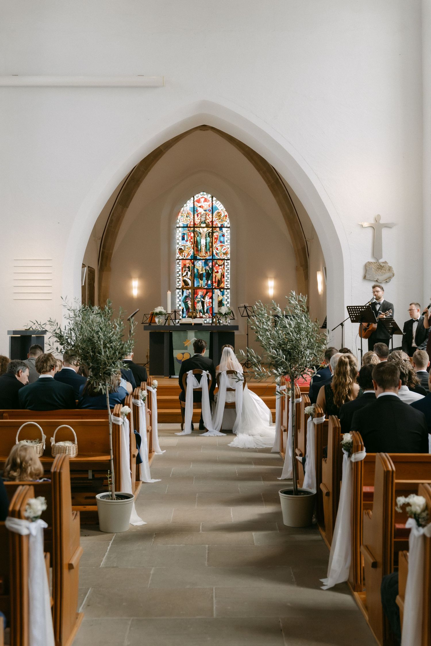 Bride and groom kneel at the altar during a church wedding ceremony, with guests seated in pews and a musician playing beside stained glass.
