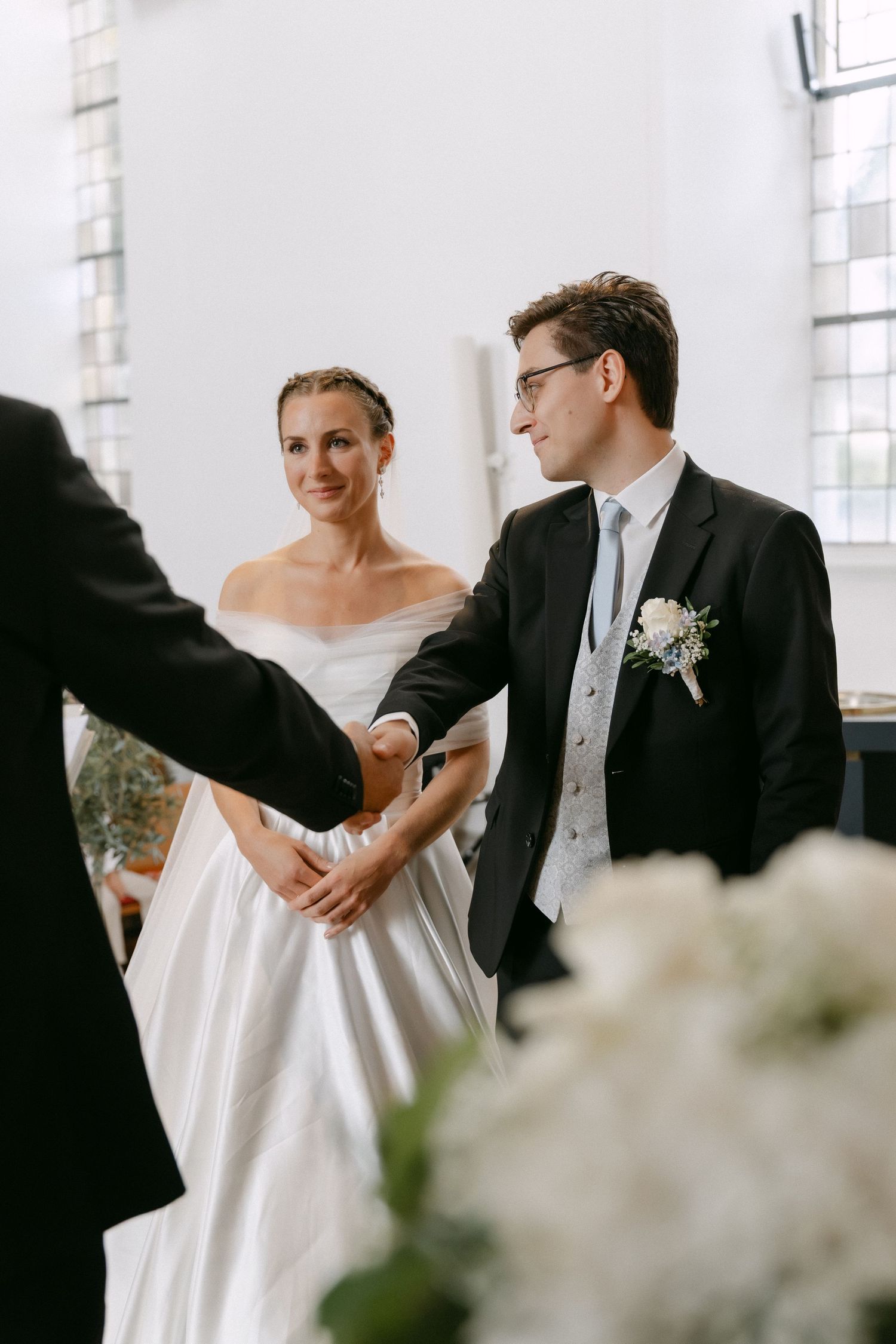 A smiling bride in an off-shoulder satin wedding gown watches as the groom in a dark suit shakes hands with a guest at the ceremony.