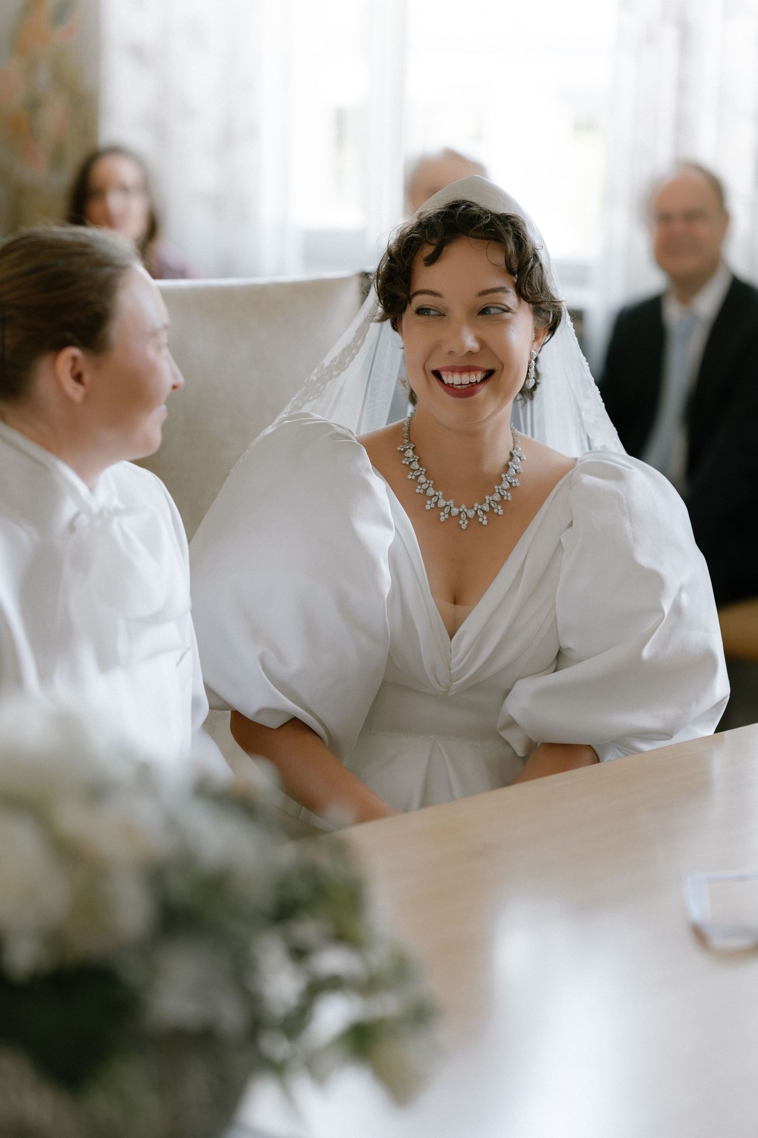 Smiling bride in white gown with puff sleeves and diamond necklace sits at table during wedding ceremony.