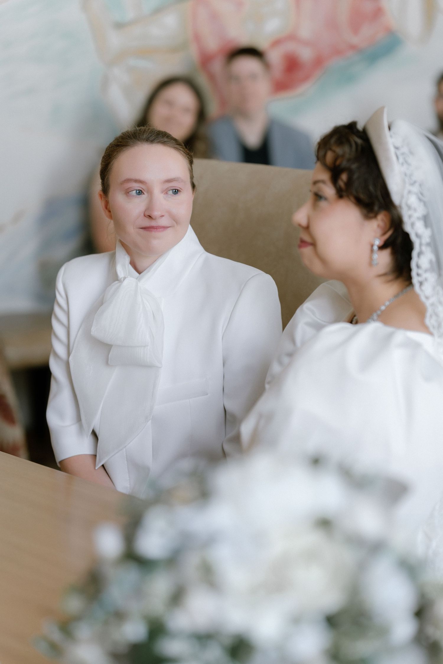 Two brides in white attire share a tender moment at their wedding ceremony, with guests visible in the background.