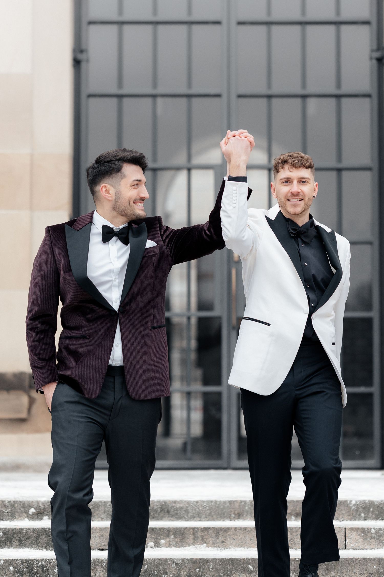 Two people in stylish tuxedos, one in burgundy and one in white, celebrate joyfully on stone steps.