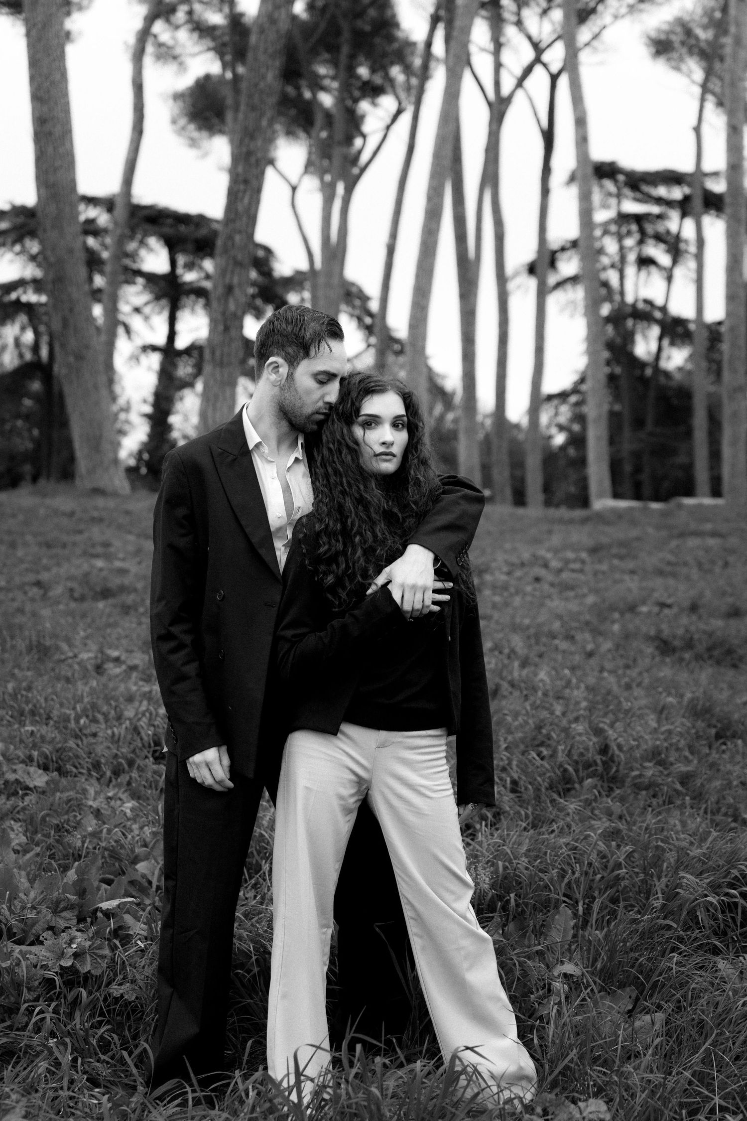 Artistic black and white portrait of a fashionable couple embracing outdoors among tall trees.