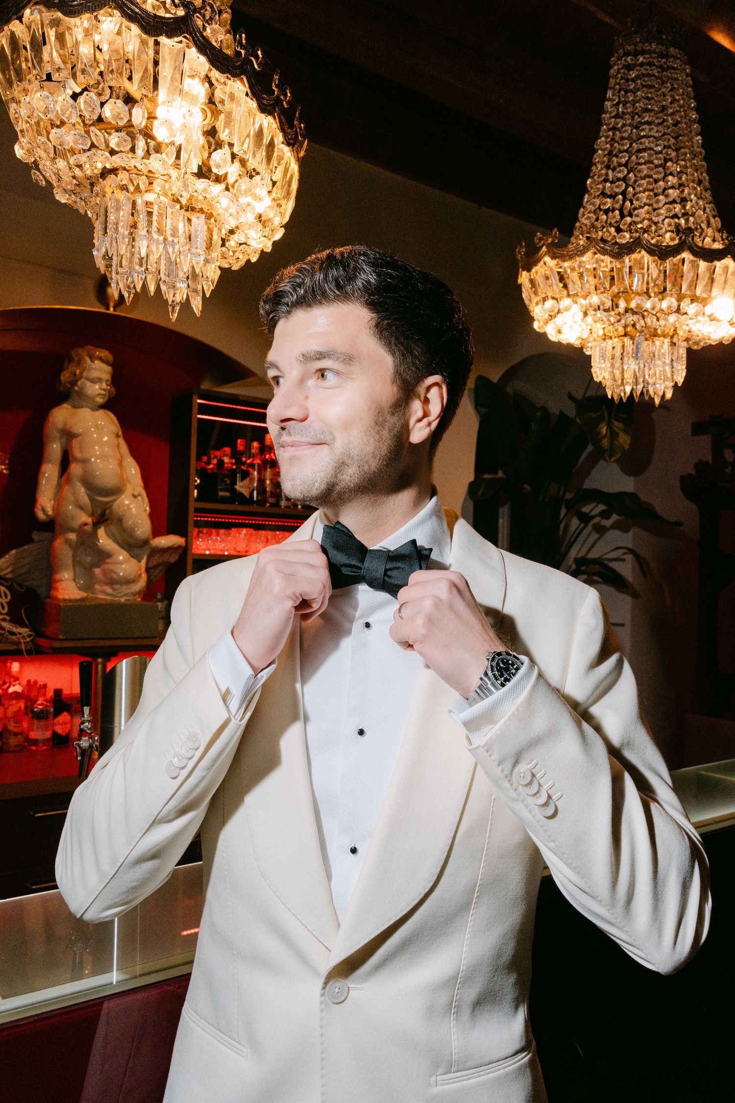 A person in a white tuxedo jacket and bow tie adjusts their collar under elegant crystal chandeliers.