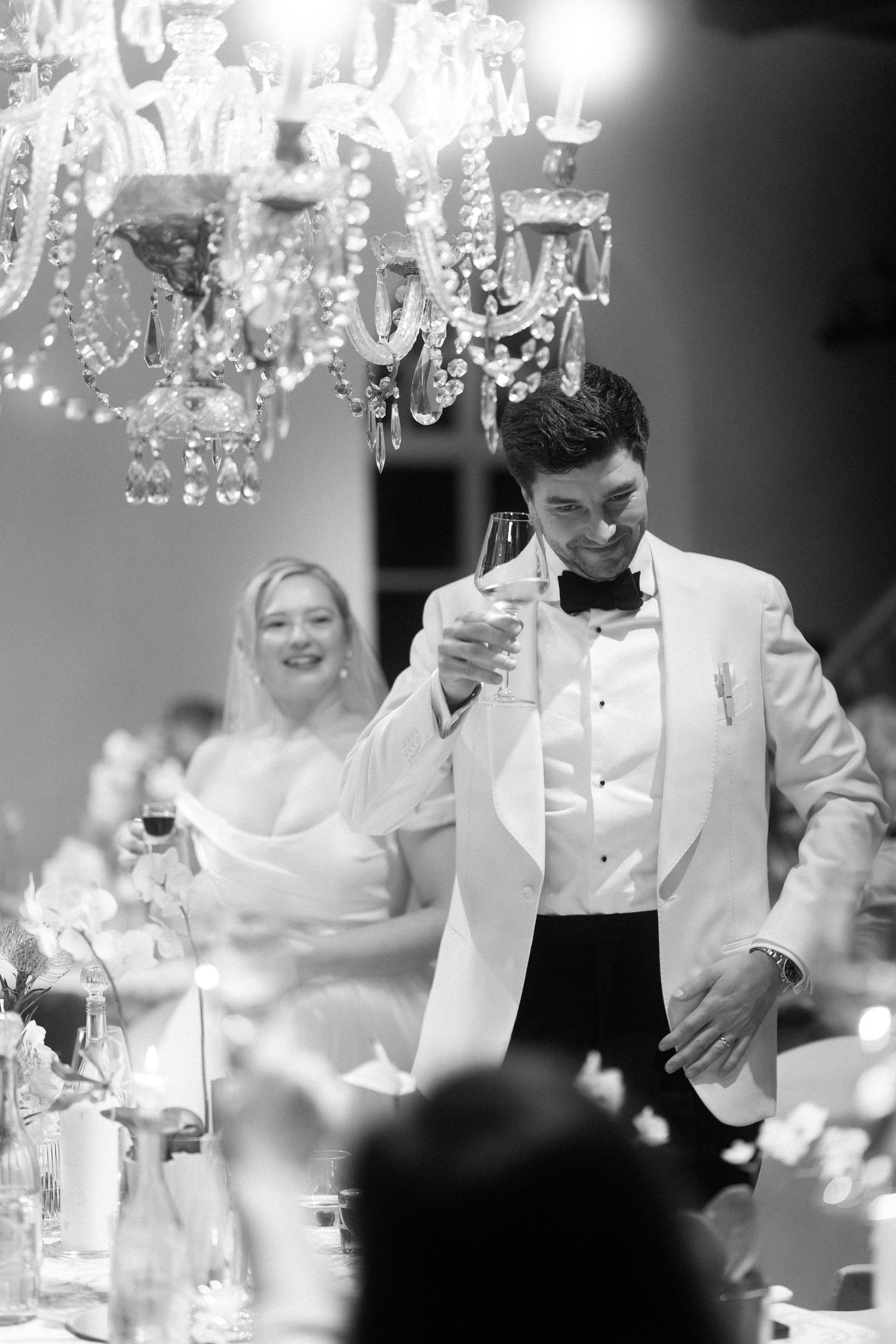 A bride and groom share a toast under an elegant crystal chandelier during their wedding reception in black and white.