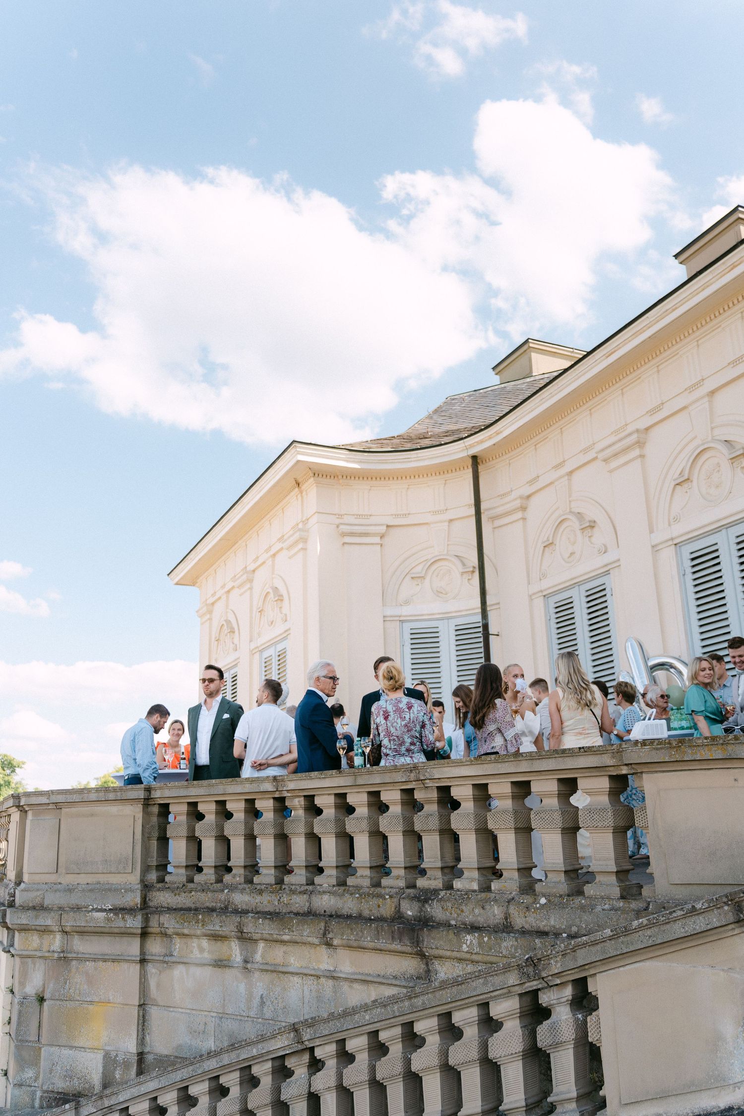 Guests gather on an elegant stone balcony of a classical building with white walls and shutters under a blue summer sky. Schloss Solitude