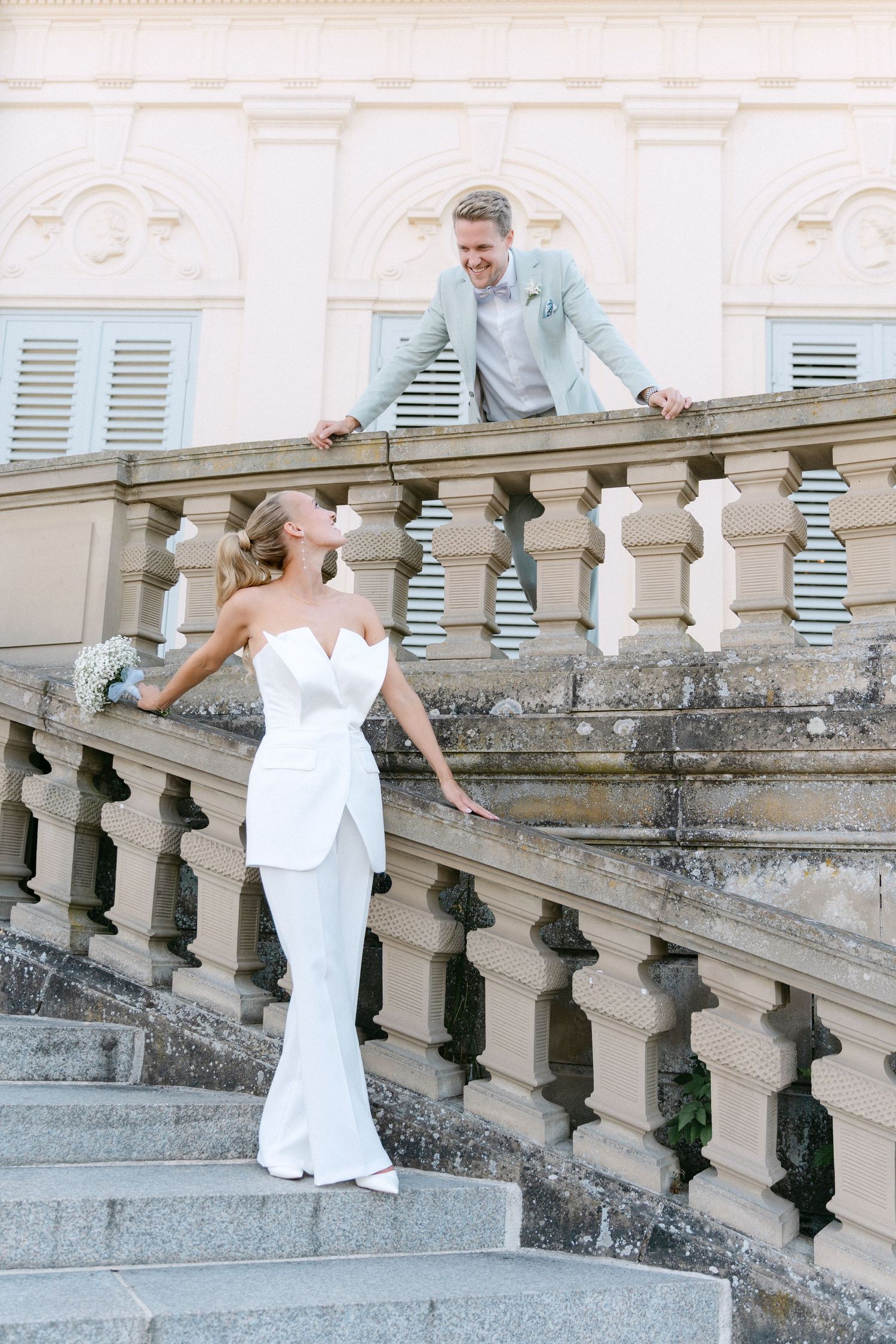 A couple in wedding attire interacts playfully on an elegant stone staircase with ornate balustrades at a historic venue. Schloss Solitude
