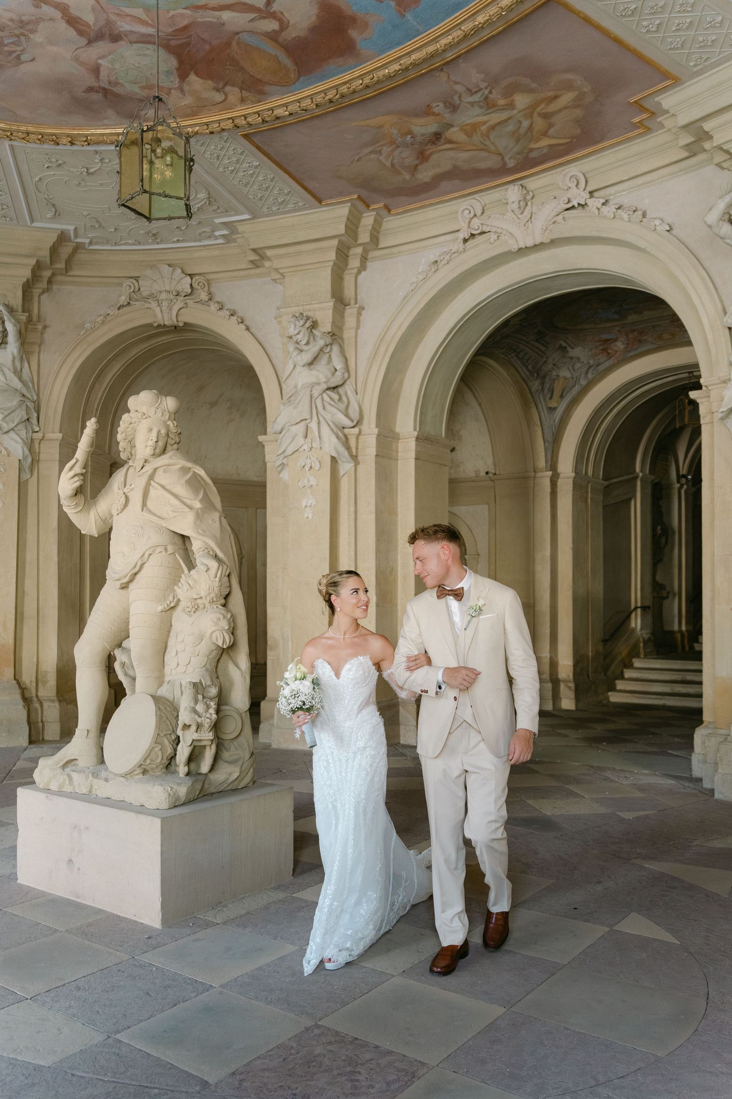 Couple walks through an ornate baroque palace corridor with white stone sculptures and painted ceiling decorations. Schloss Ludwigsburg