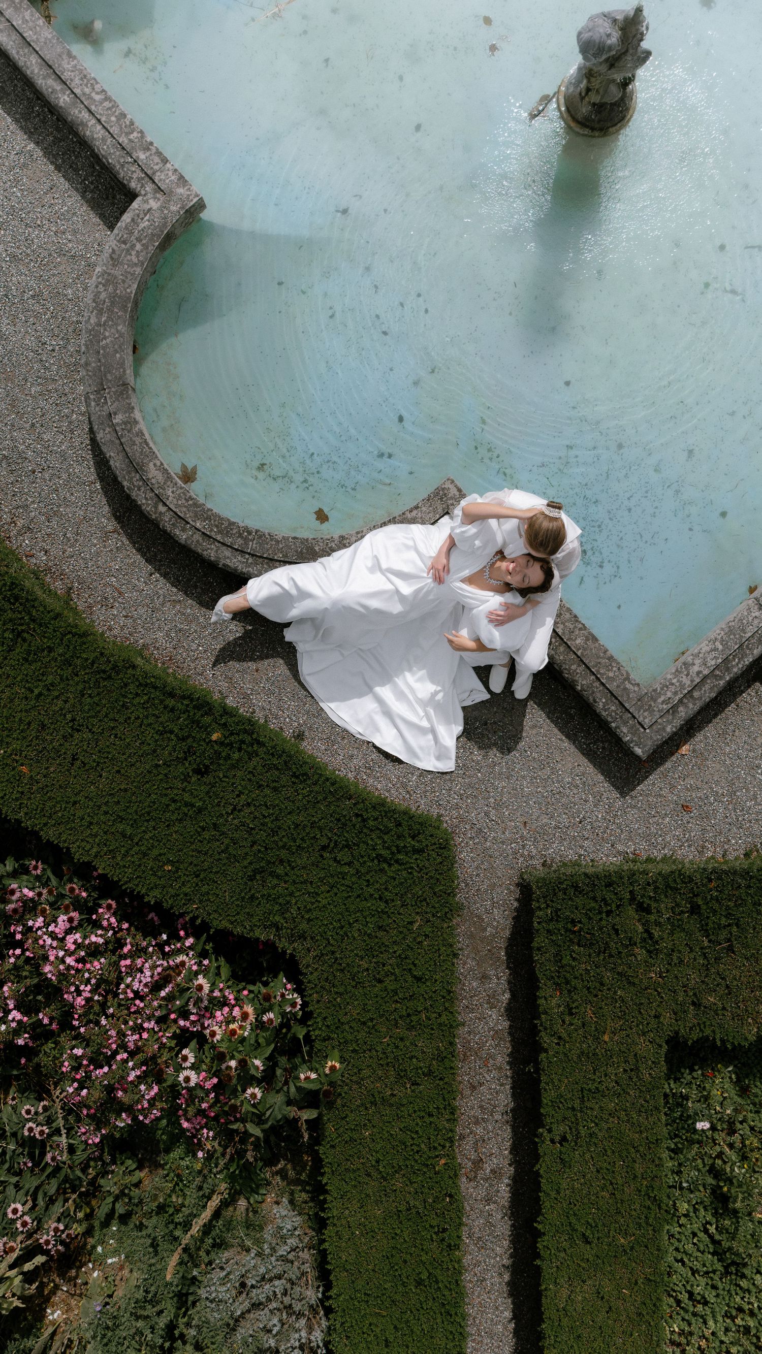Aerial view of a couple in white wedding attire embracing beside an ornate garden fountain with manicured hedges.