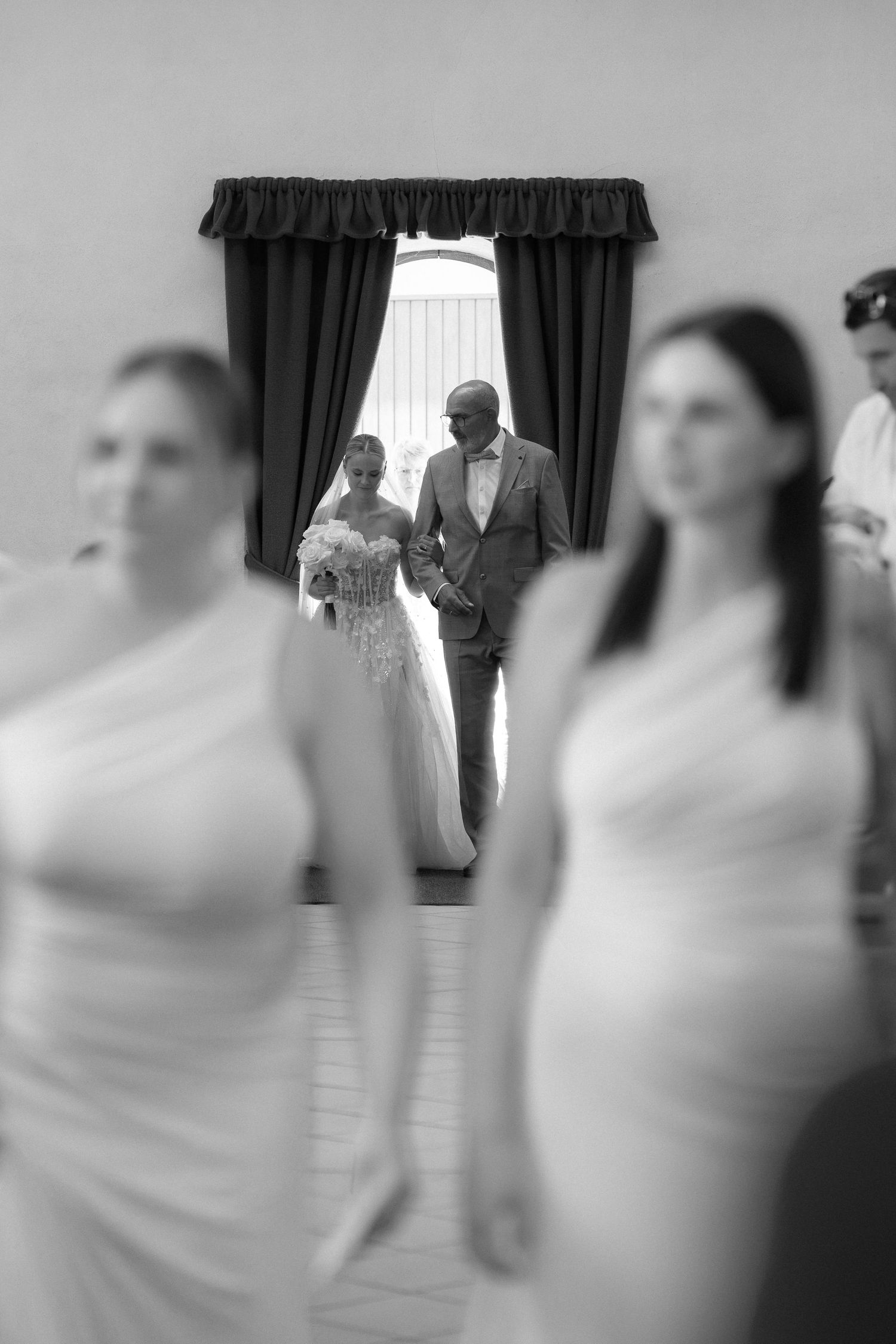 Black and white wedding photo of a bride in a lace gown walking with an older man in a suit, framed by curtains behind blurred bridesmaids.