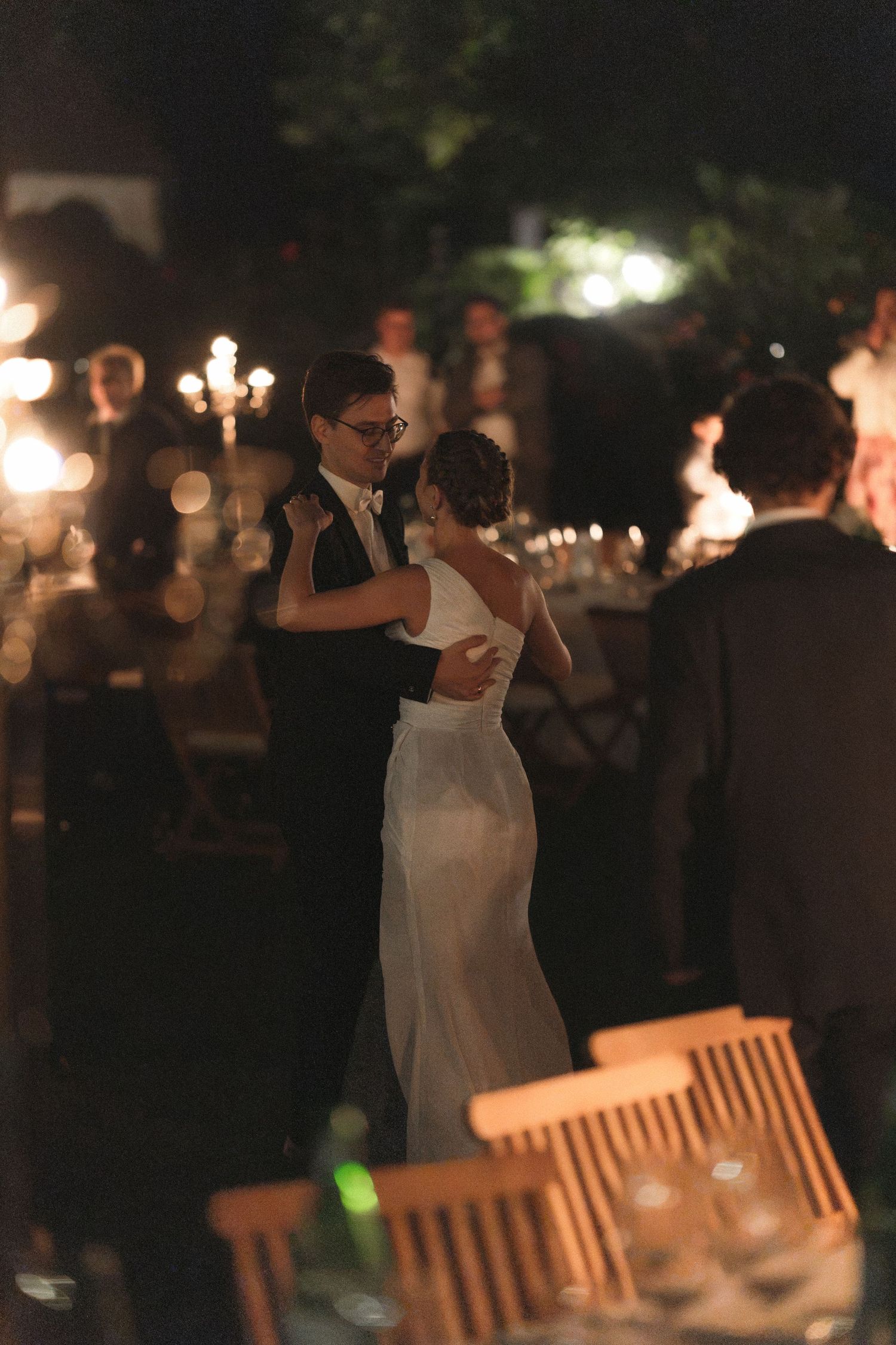 Bride in a white gown and groom in a dark suit share a romantic first dance at an outdoor evening wedding reception with candlelit tables.