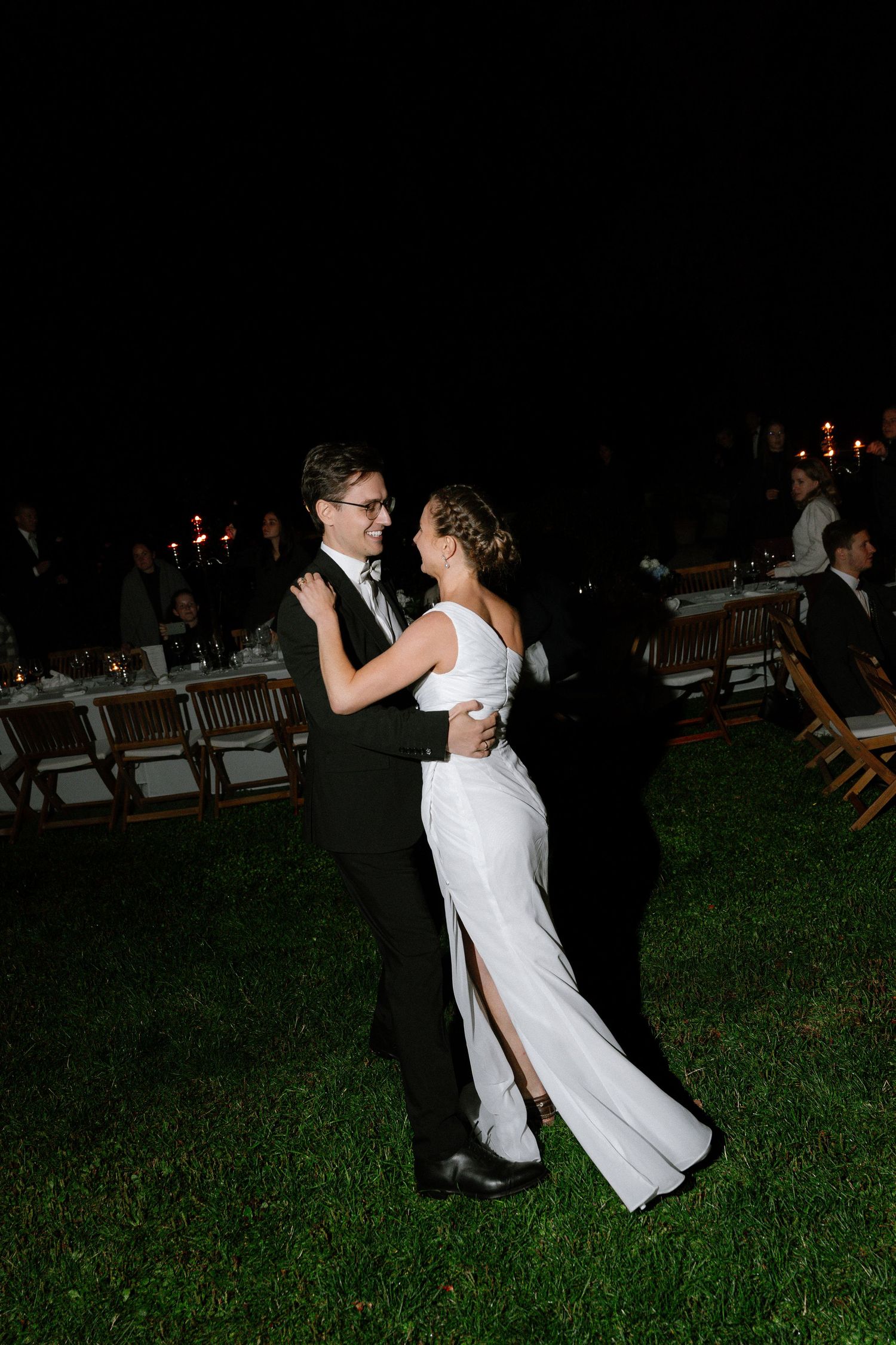 Couple in formal wear dance on a grass lawn at night during an outdoor wedding reception with candlelit tables and guests behind them.