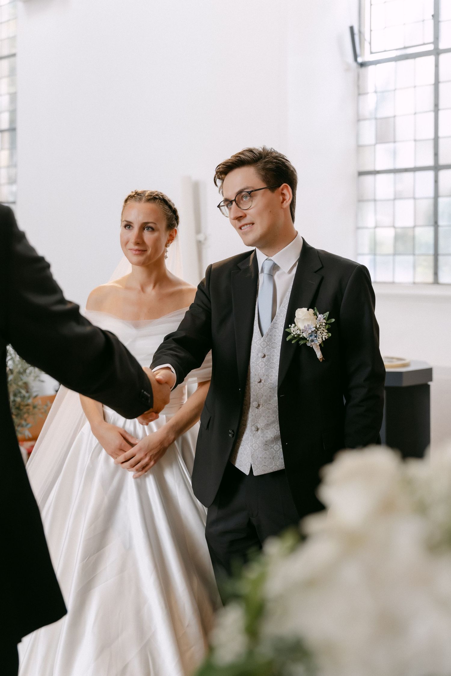 Bride in a white satin gown and groom in a dark suit with floral boutonniere and glasses hold hands during a bright indoor wedding ceremony.