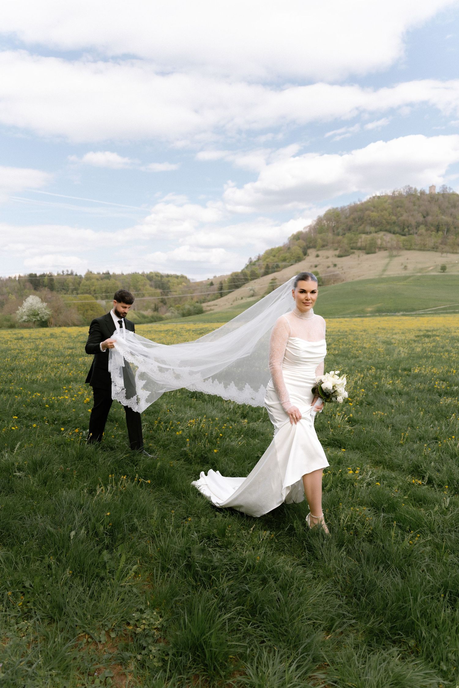 Bride in white gown with flowing veil walks through green meadow as groom in black suit holds veil on sunny day.