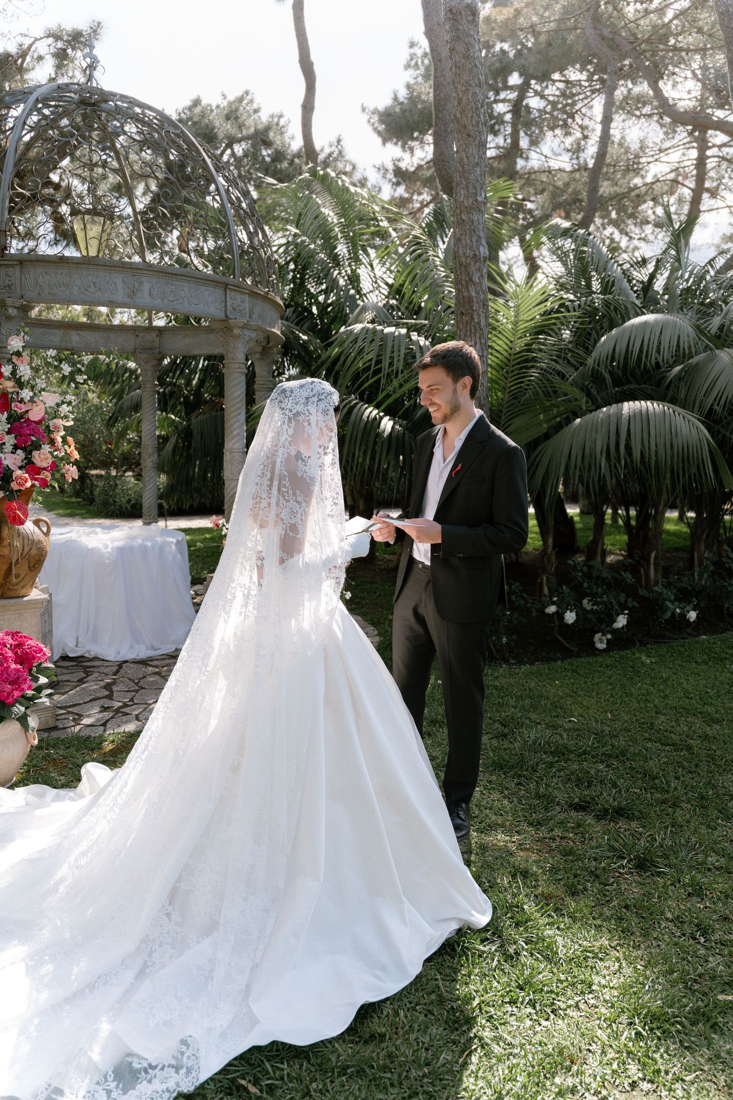 Groom reads vows to bride in white lace gown and veil during outdoor garden wedding ceremony.