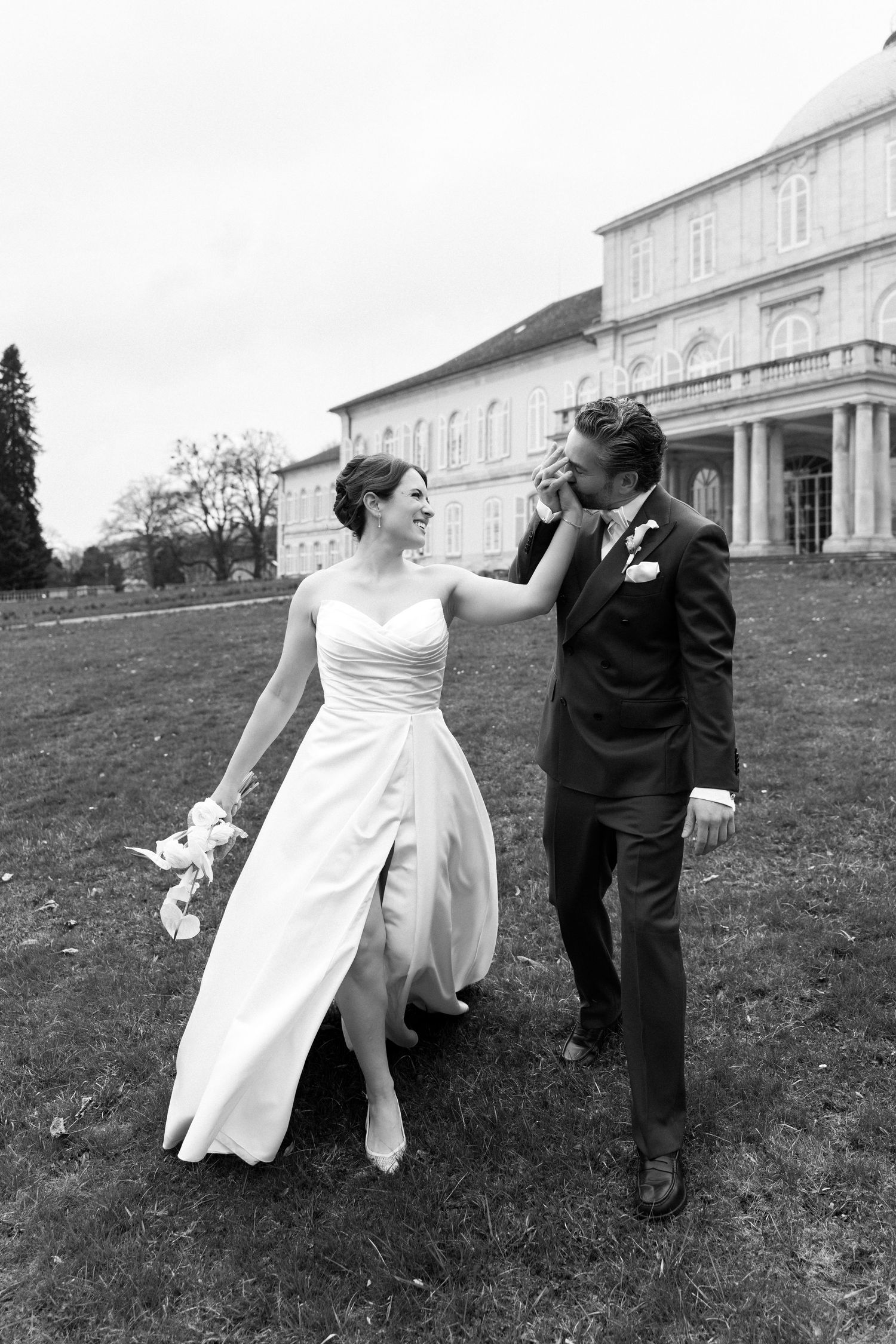 Black and white photo of a smiling bride in a white gown and groom in a suit walking outside a grand estate.
