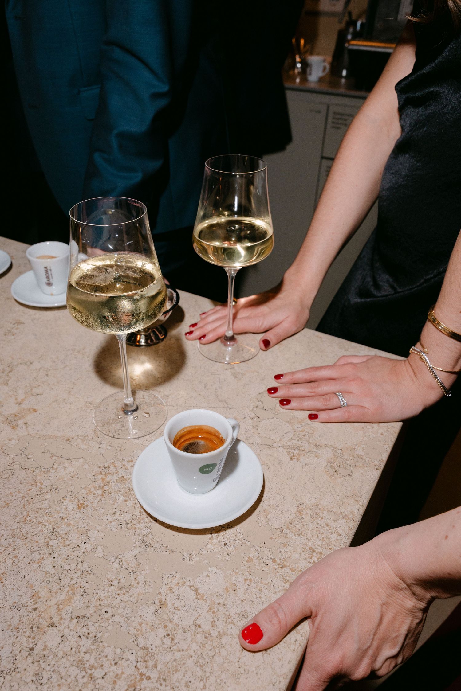 Elegant party scene with two white wine glasses and espresso cup on marble counter, with guests in formal attire.