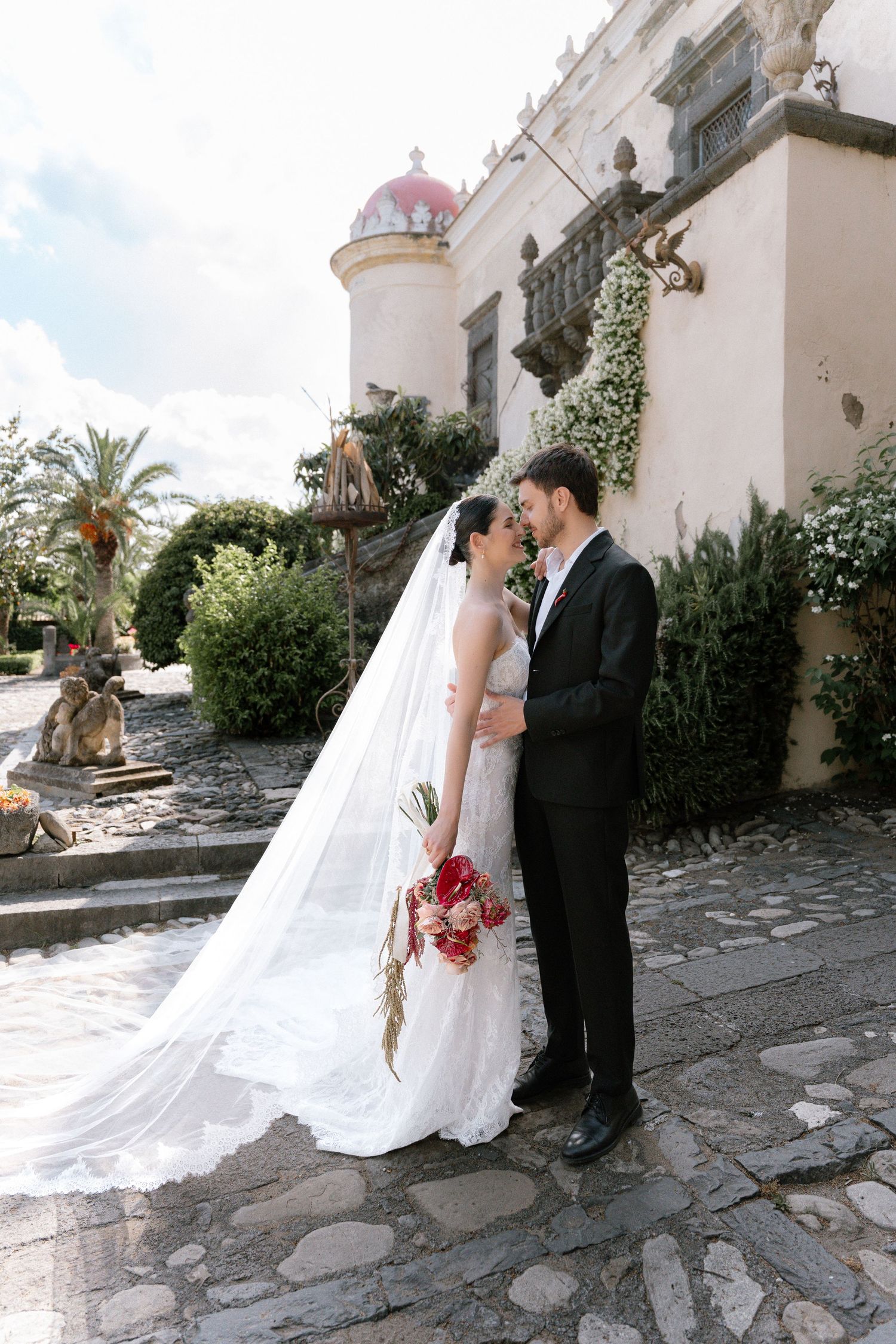 Bride and groom share a romantic moment outside a historic stone building, bride holding a red bouquet with long veil flowing.