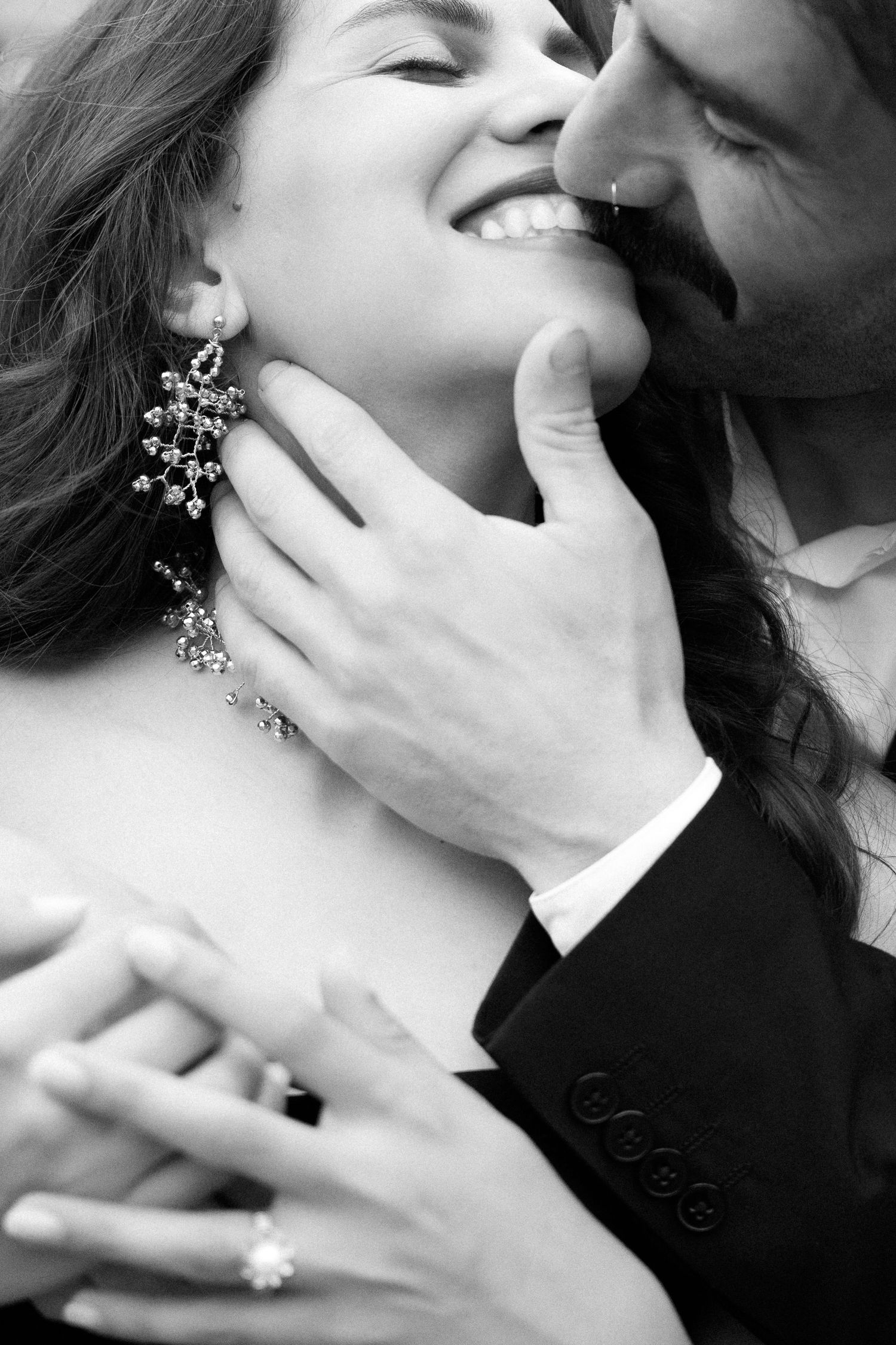 Black and white photo of a smiling bride wearing diamond earrings and ring, embraced by groom in suit.