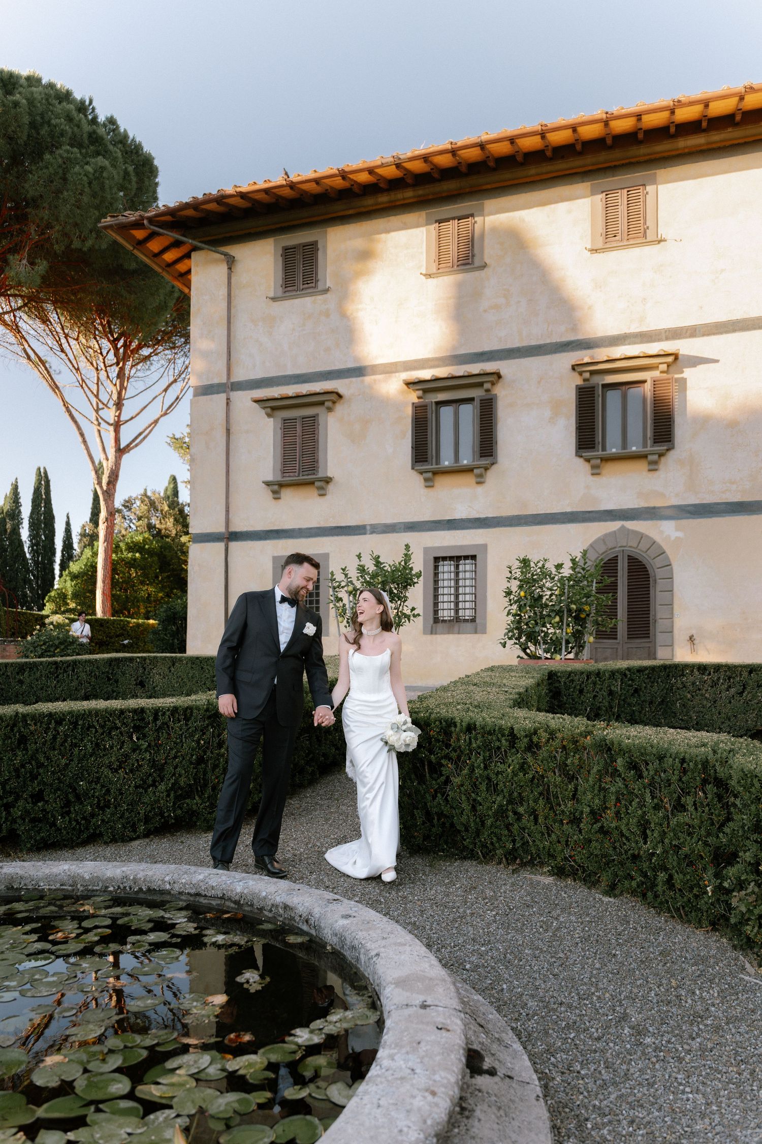 Bride and groom walk hand in hand beside a lily pond in a manicured Italian villa garden at golden hour.