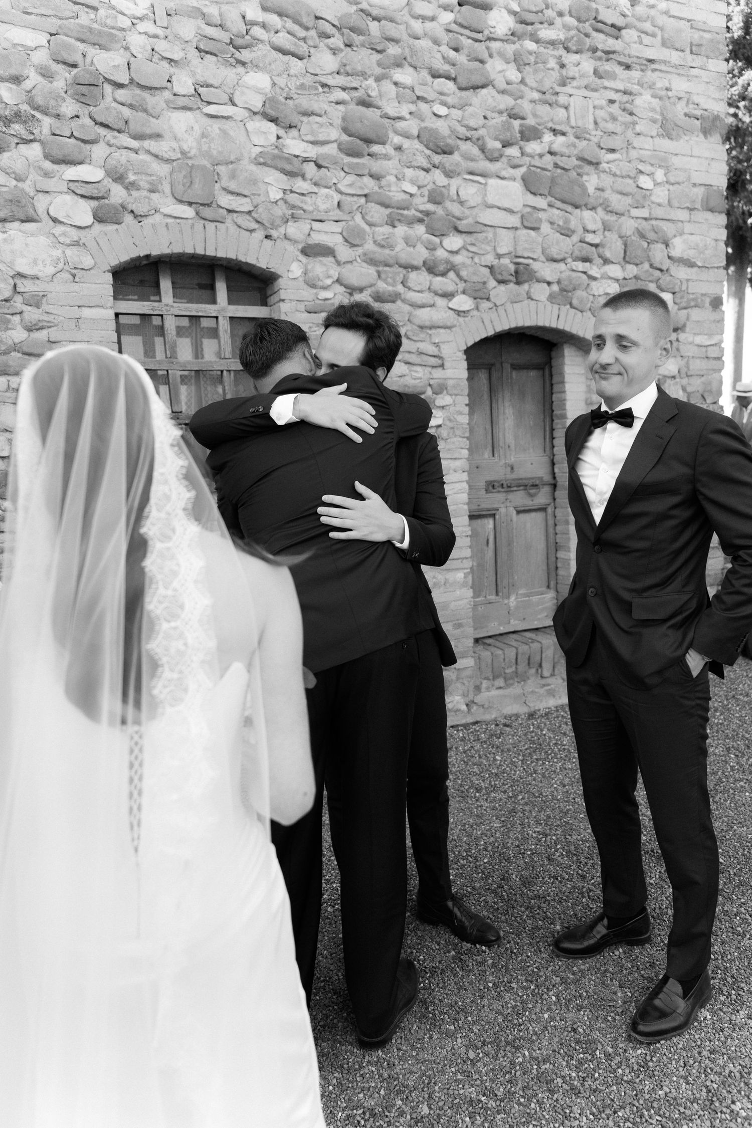 Black and white wedding photo of bride and groom embracing near stone building while guest smiles nearby.