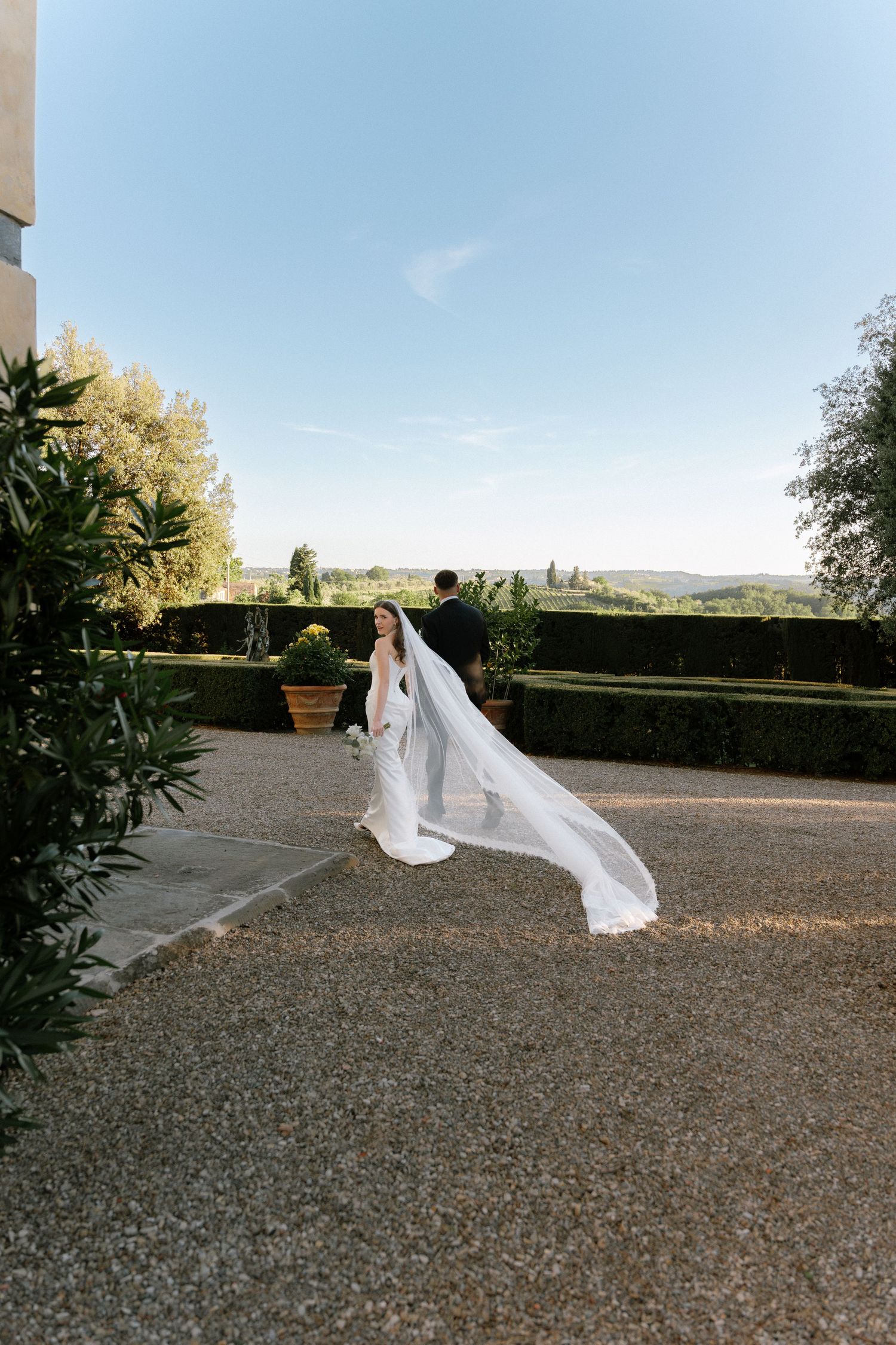 Bride in flowing white gown and veil blowing in wind stands in elegant garden courtyard with groom and scenic views.