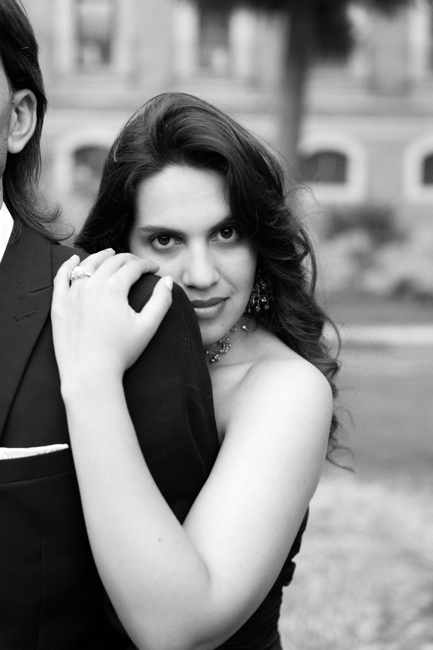 Black and white portrait of elegant woman in black dress with jewelry, holding man's arm outdoors.