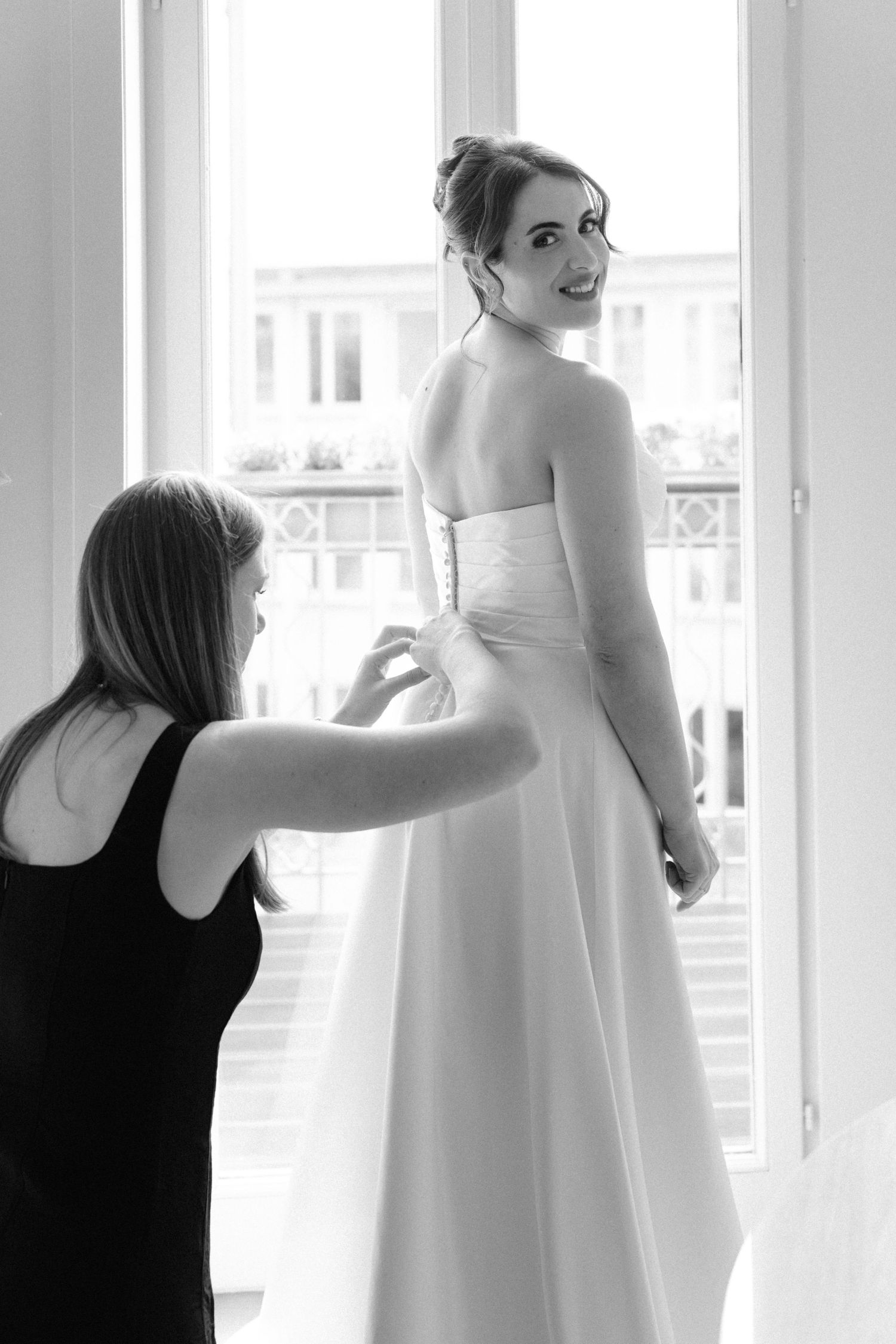 Black and white photo of a bride in a white gown being helped by a bridesmaid near a bright window.