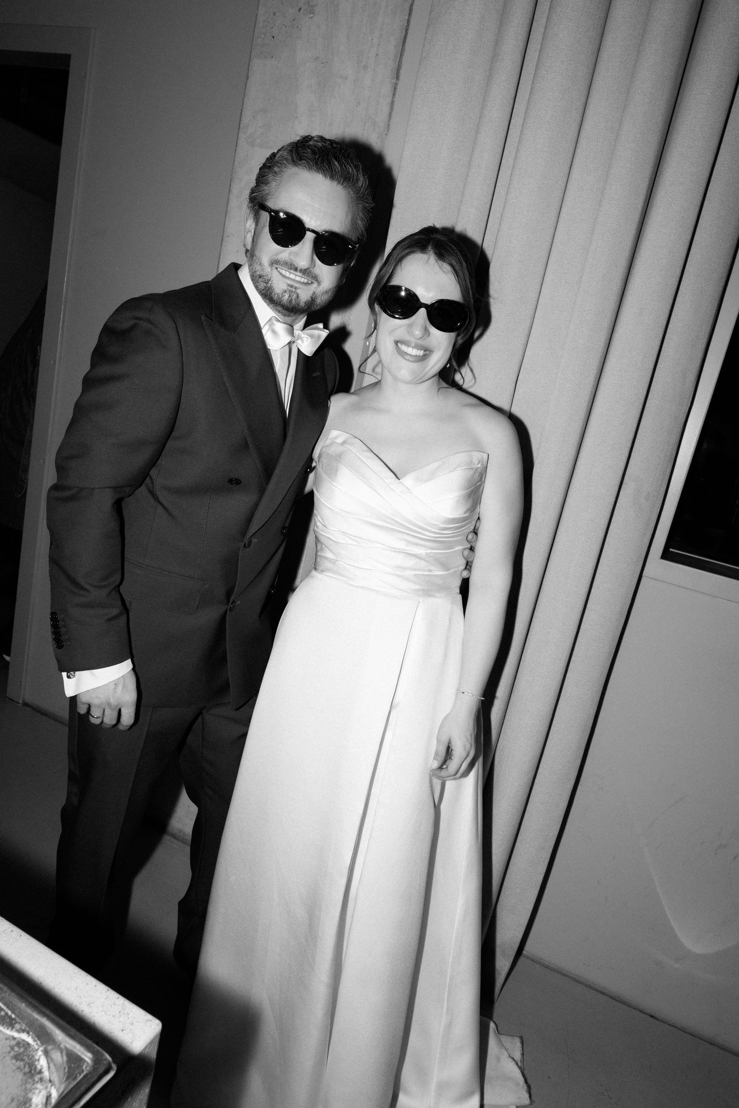 Black and white photo of a smiling bride in white gown and groom in suit, both wearing sunglasses at wedding reception.