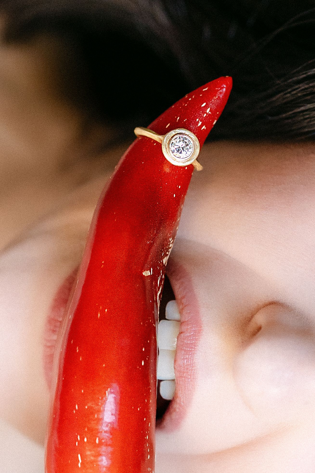 A gold bezel-set diamond ring displayed on a red chili pepper held between a woman's teeth.
