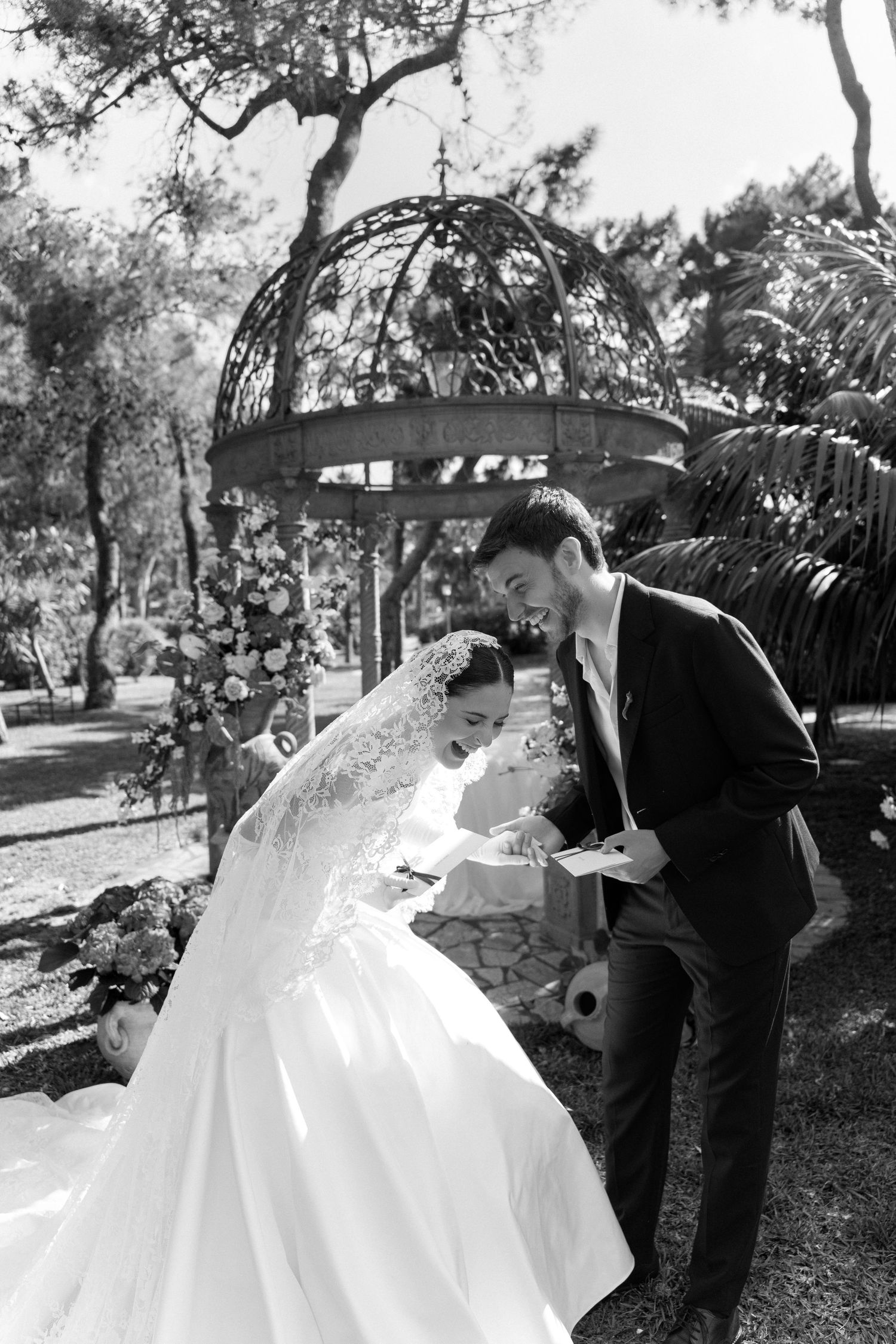 Black and white photo of bride and groom signing wedding documents outdoors near floral arch and ornate gazebo.