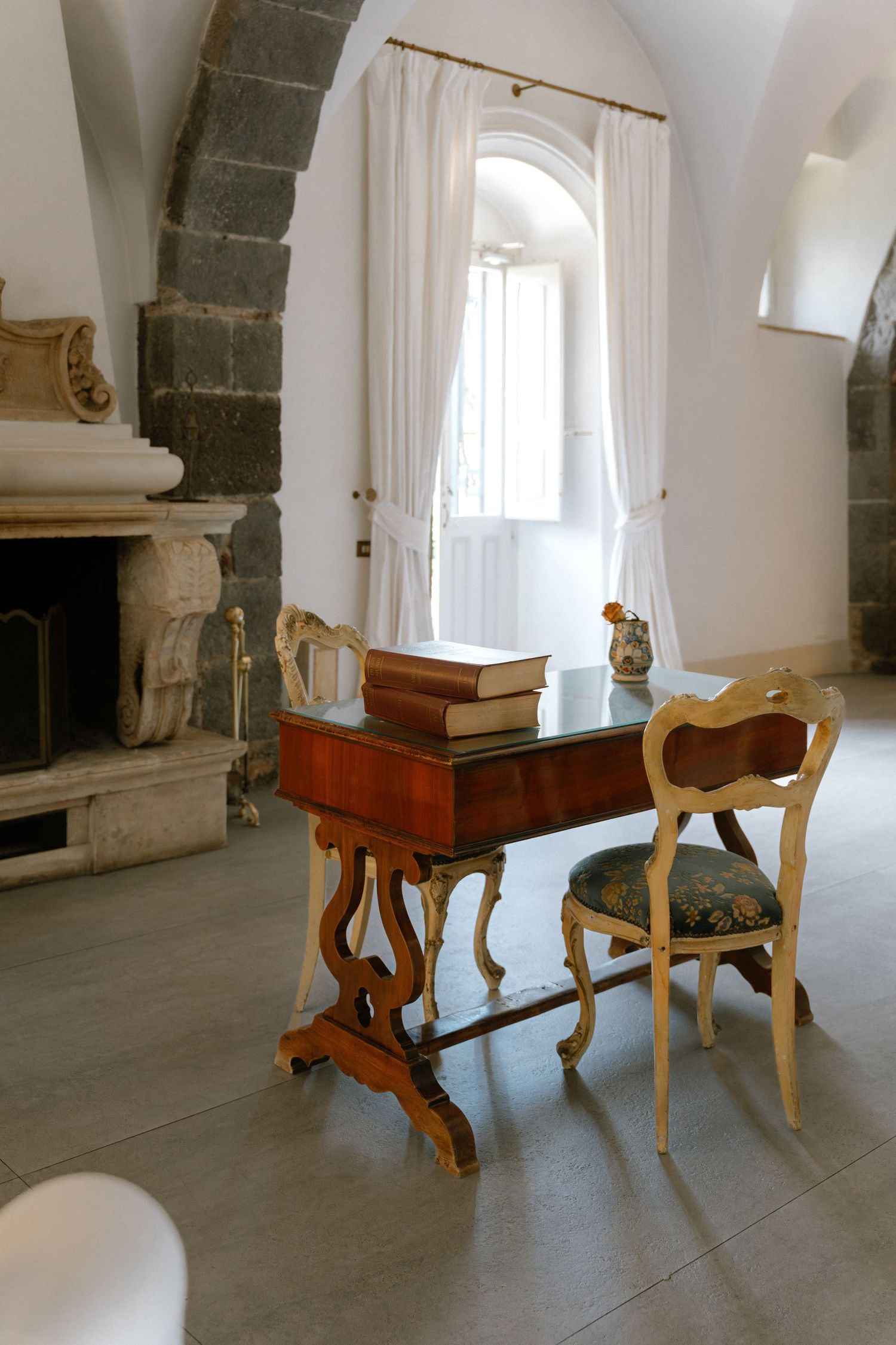 Elegant antique wooden desk and ornate chair in a historic room with arched window and stone fireplace.