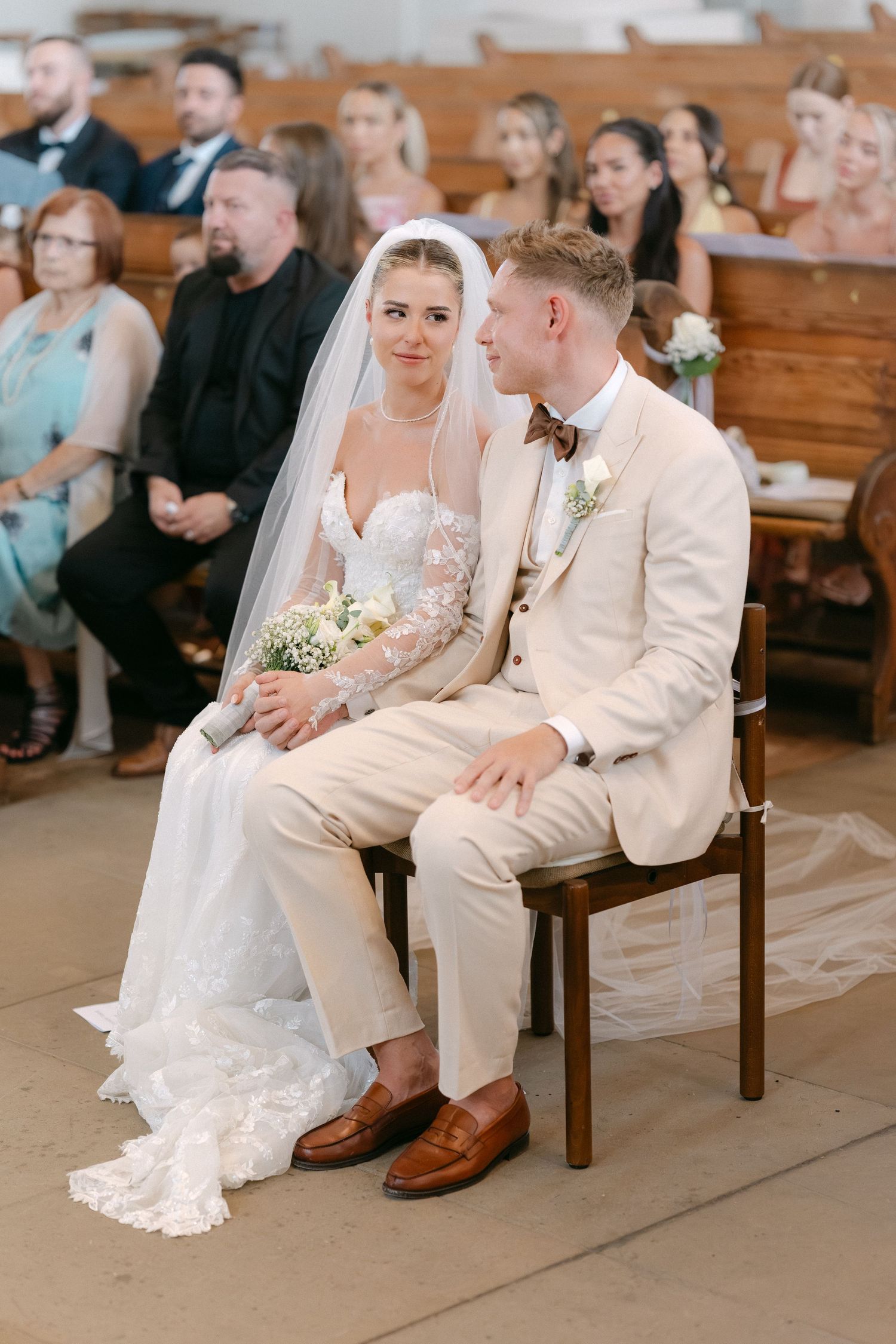 A couple sits together in a church pew during their wedding ceremony wearing white and cream colored attire.