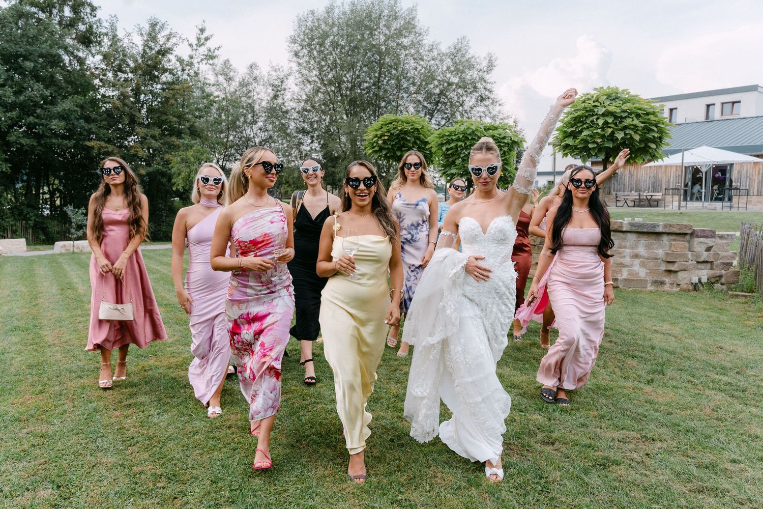 A group in flowing pink and yellow dresses walk together across a grassy lawn during a summer celebration.
