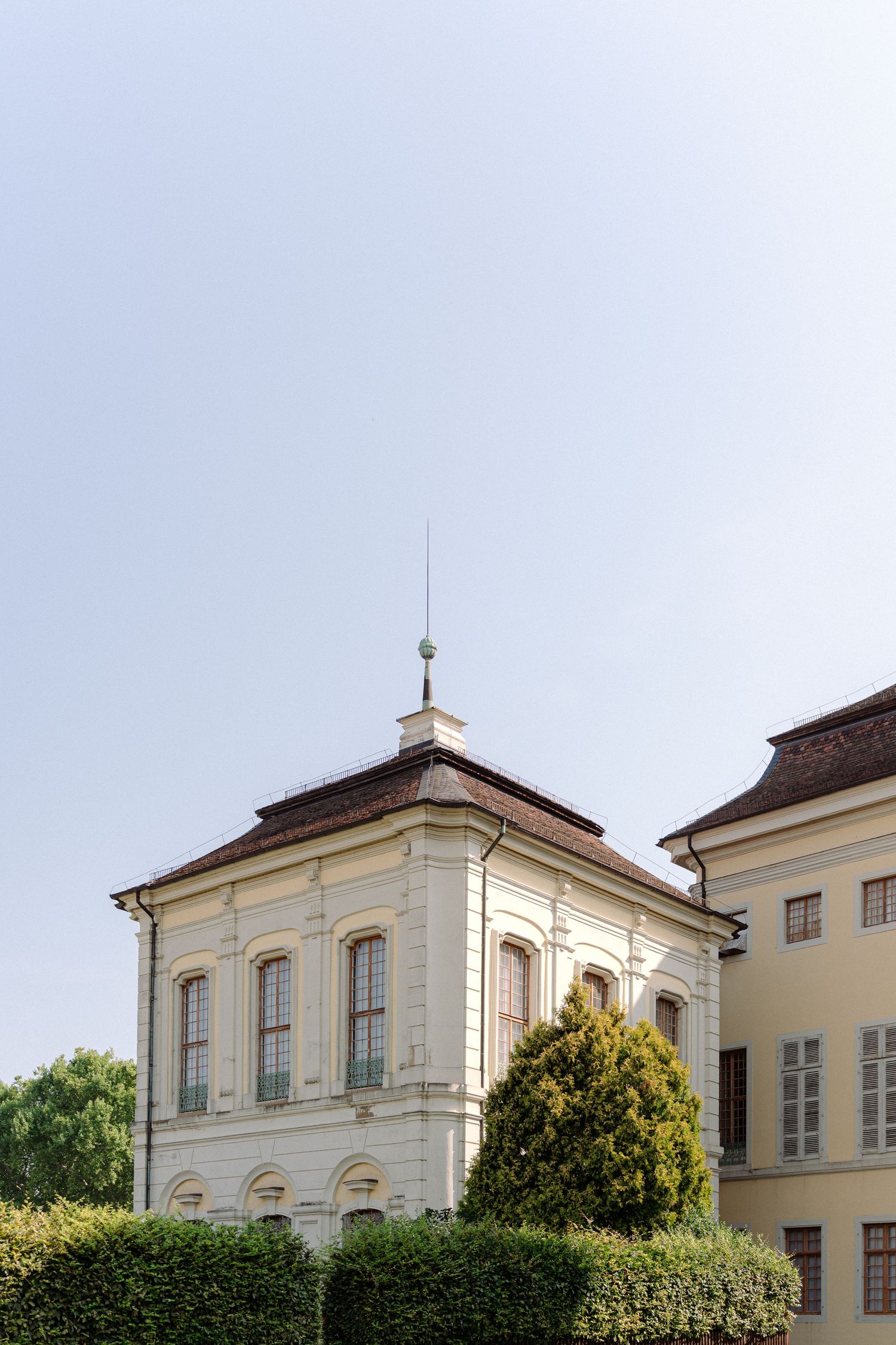 Classical white building with tall windows and ornate architecture sits among manicured hedges against a pale blue sky. Schloss Ludwigsburg