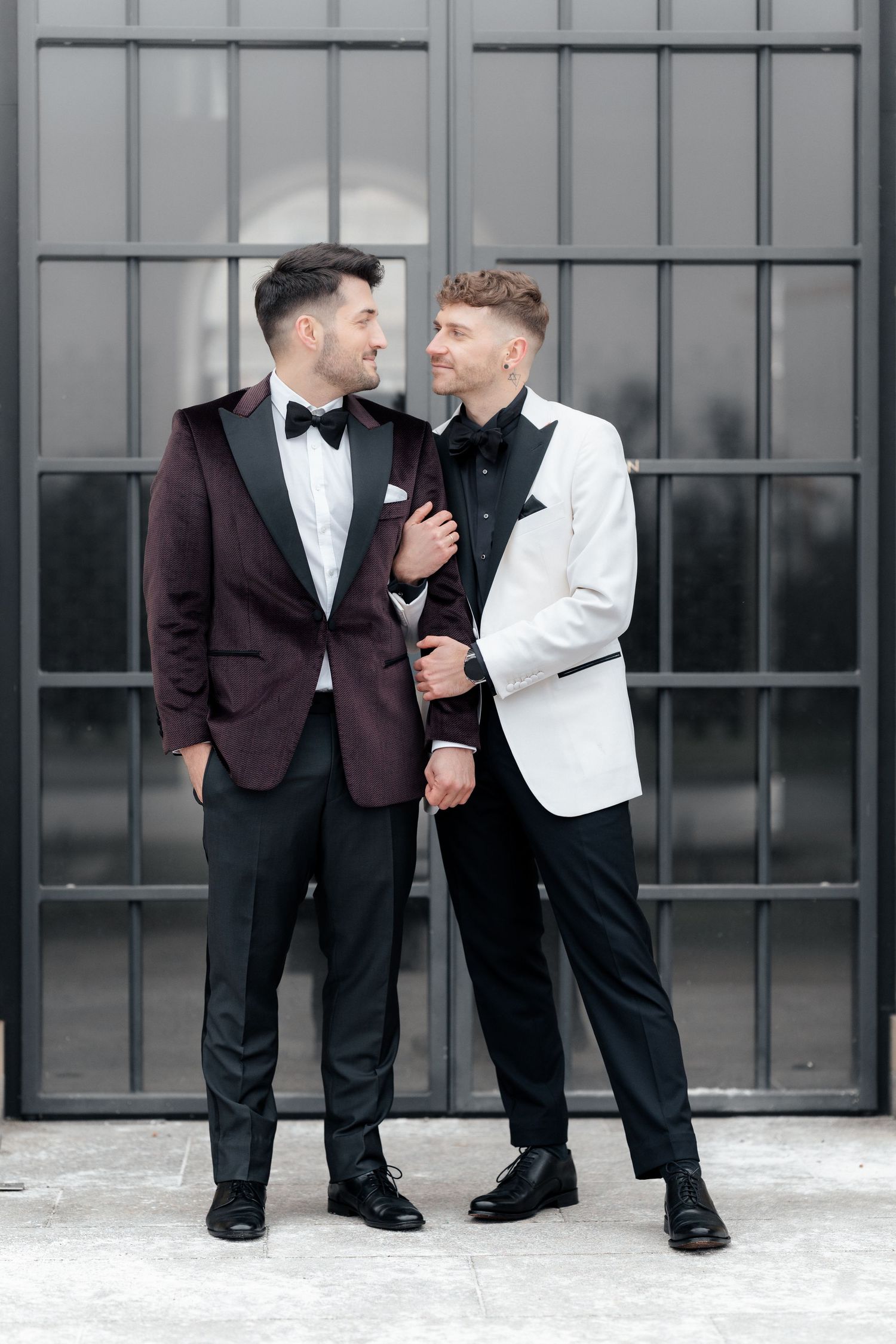 Two people in formal tuxedos share an intimate moment while posing against an industrial metal door backdrop.