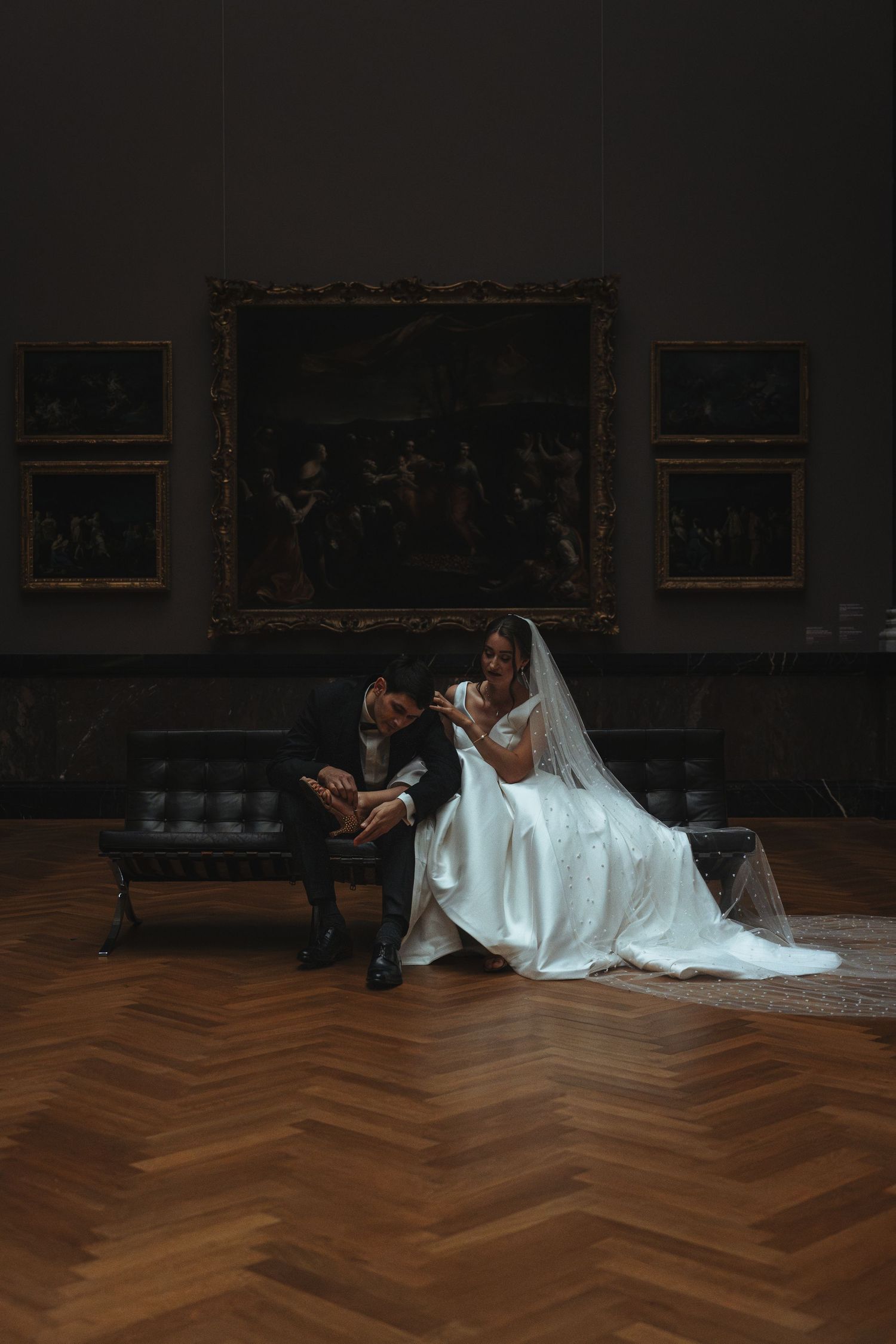 A couple in wedding attire sits on a leather couch in an art gallery with ornate frames on dark walls and herringbone floors.