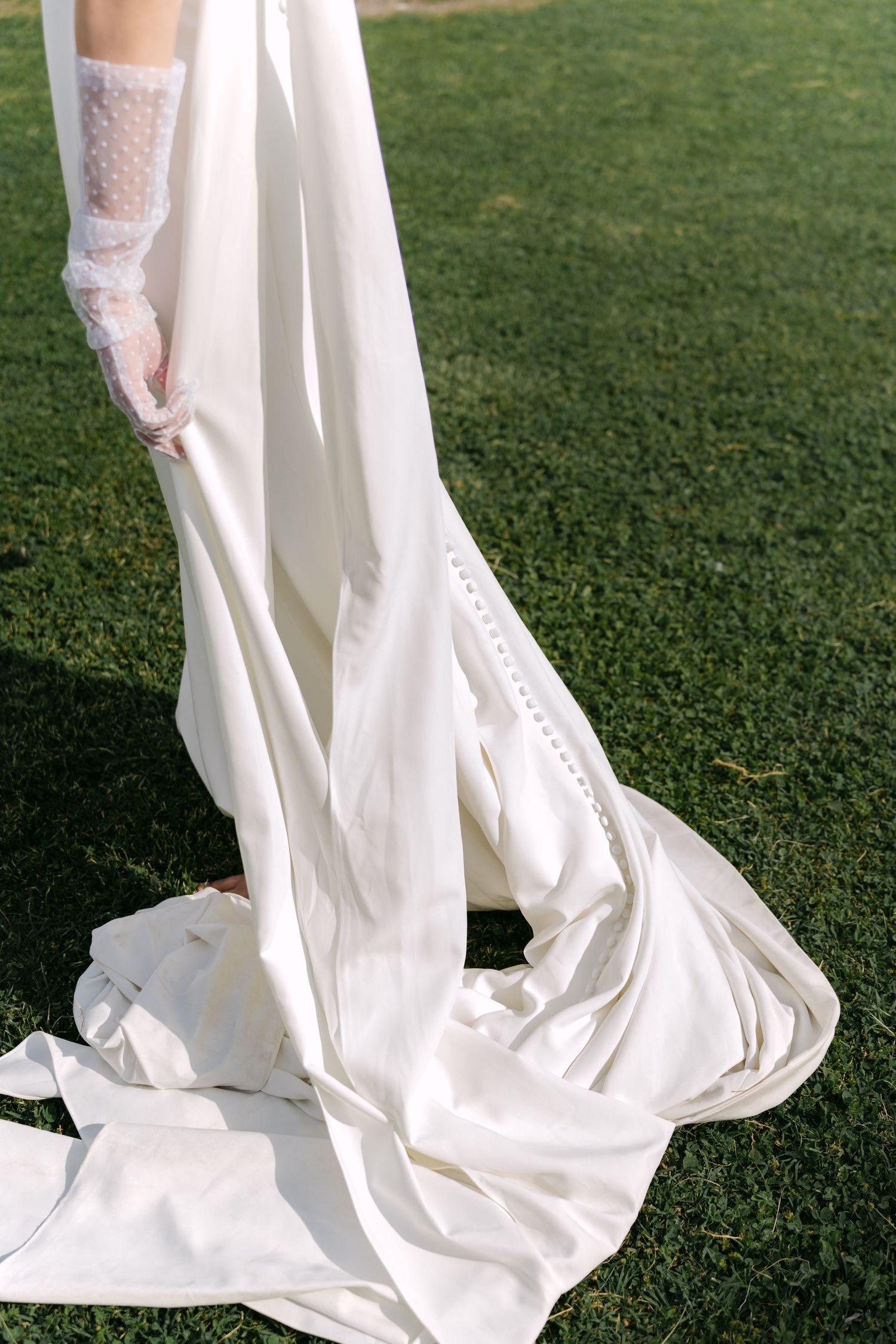 Close-up of a white lace wedding dress train flowing elegantly across green grass outdoors. Villa Mosconi Bertani