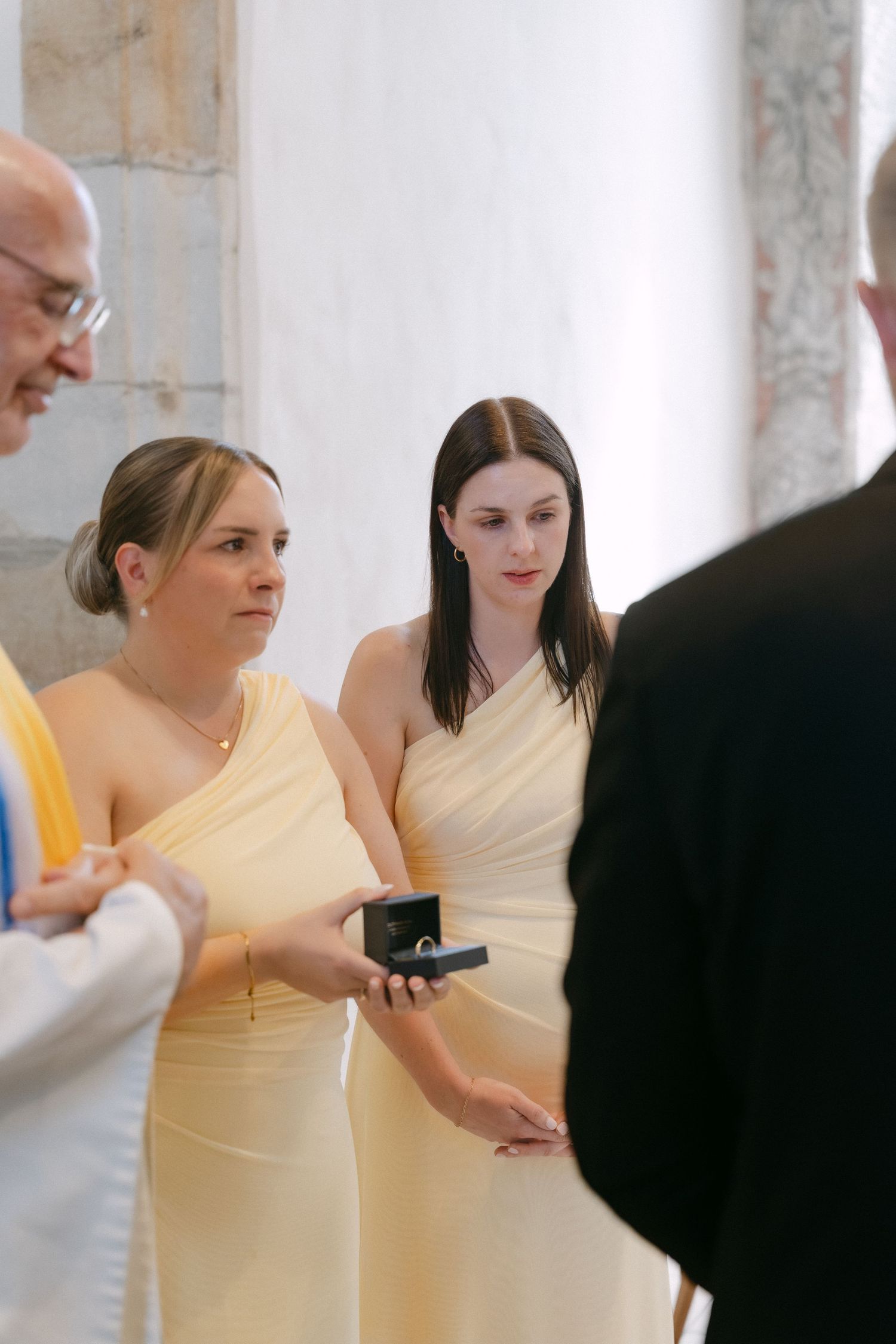 Two bridesmaids in pale yellow dresses stand during a wedding ceremony as one holds an open ring box beside an officiant.