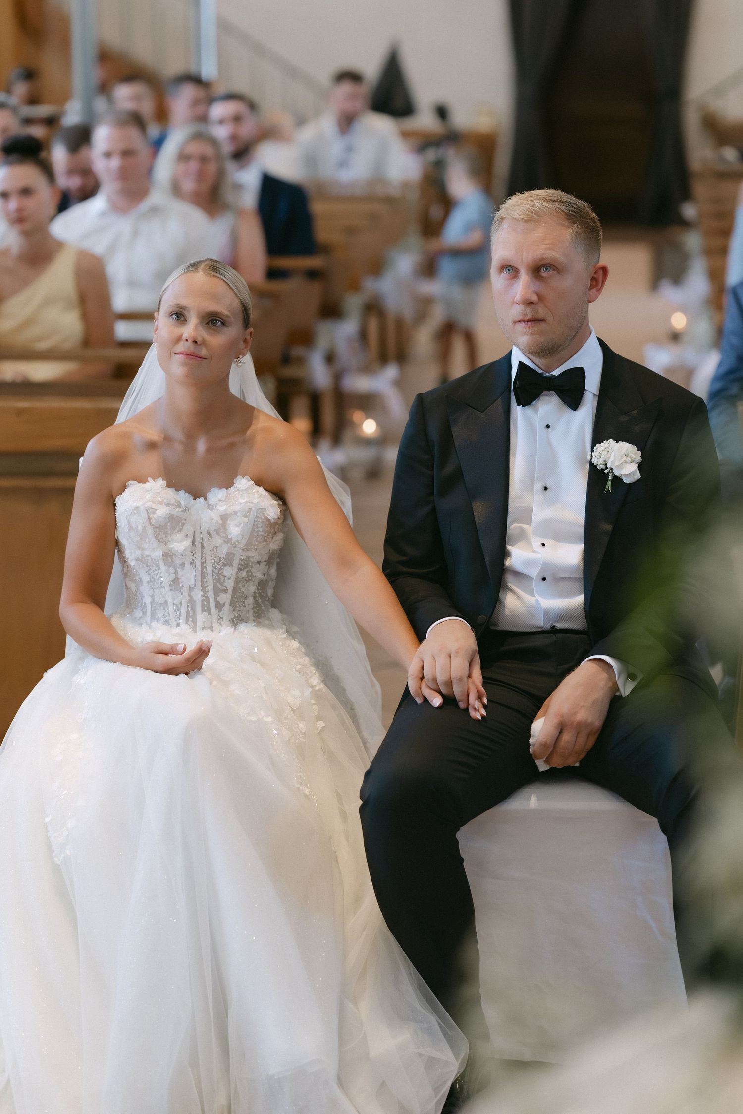Bride in a strapless lace wedding gown and veil sits beside a groom in a black tuxedo and bow tie during a church ceremony.