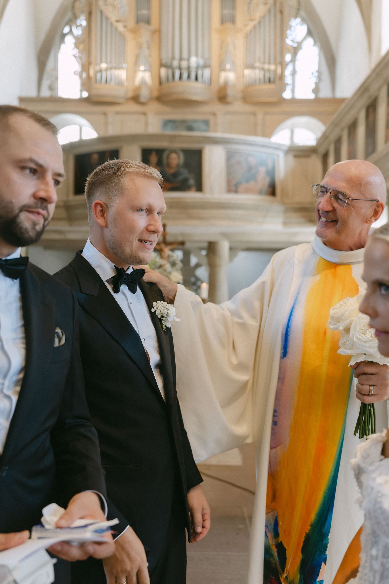 Smiling groom in a black tuxedo with a floral boutonniere stands beside a priest in colorful vestments during a church wedding ceremony.