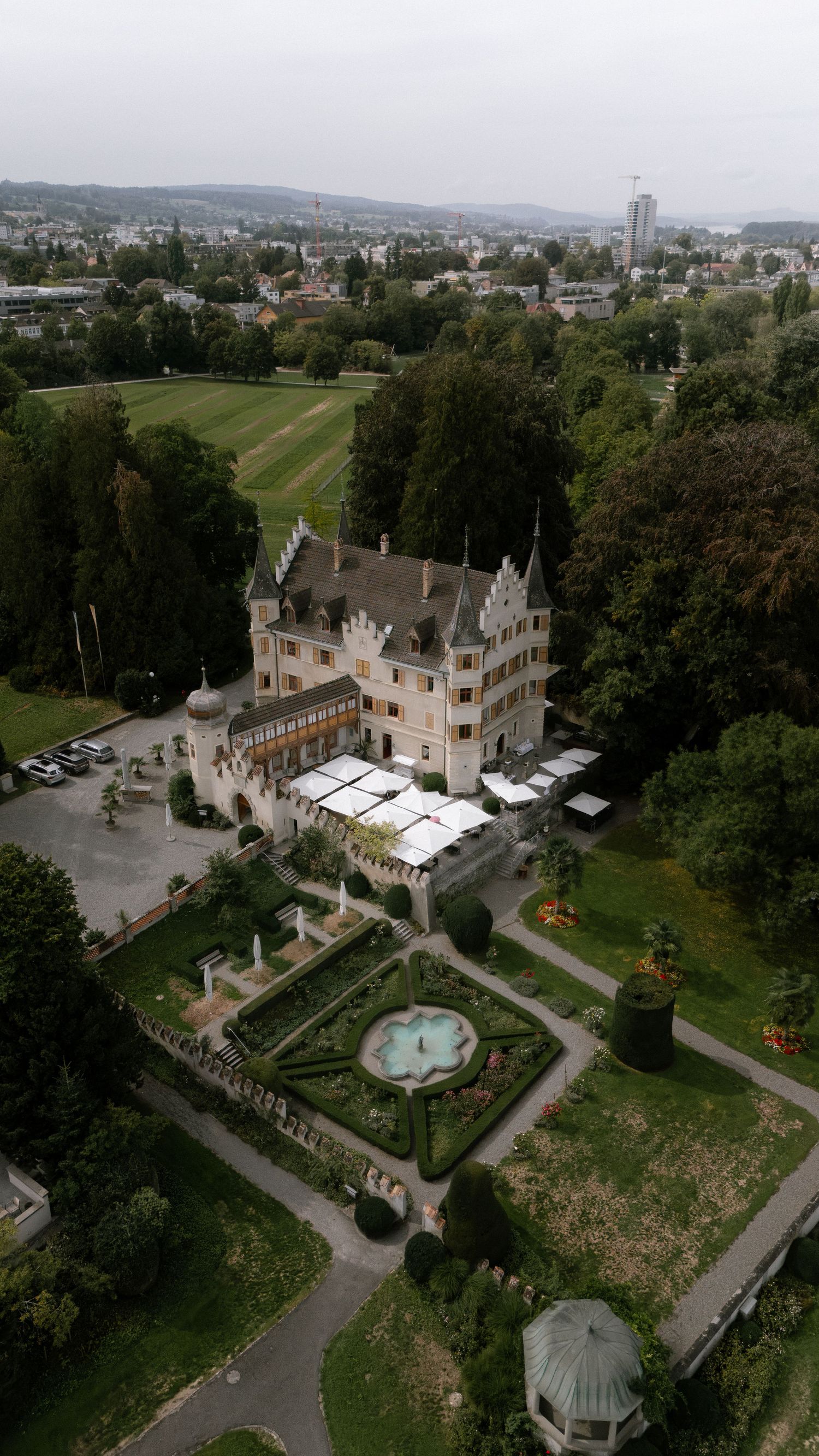 Aerial view of a historic European castle with formal gardens, fountain, and surrounding lush greenery in an urban setting.
