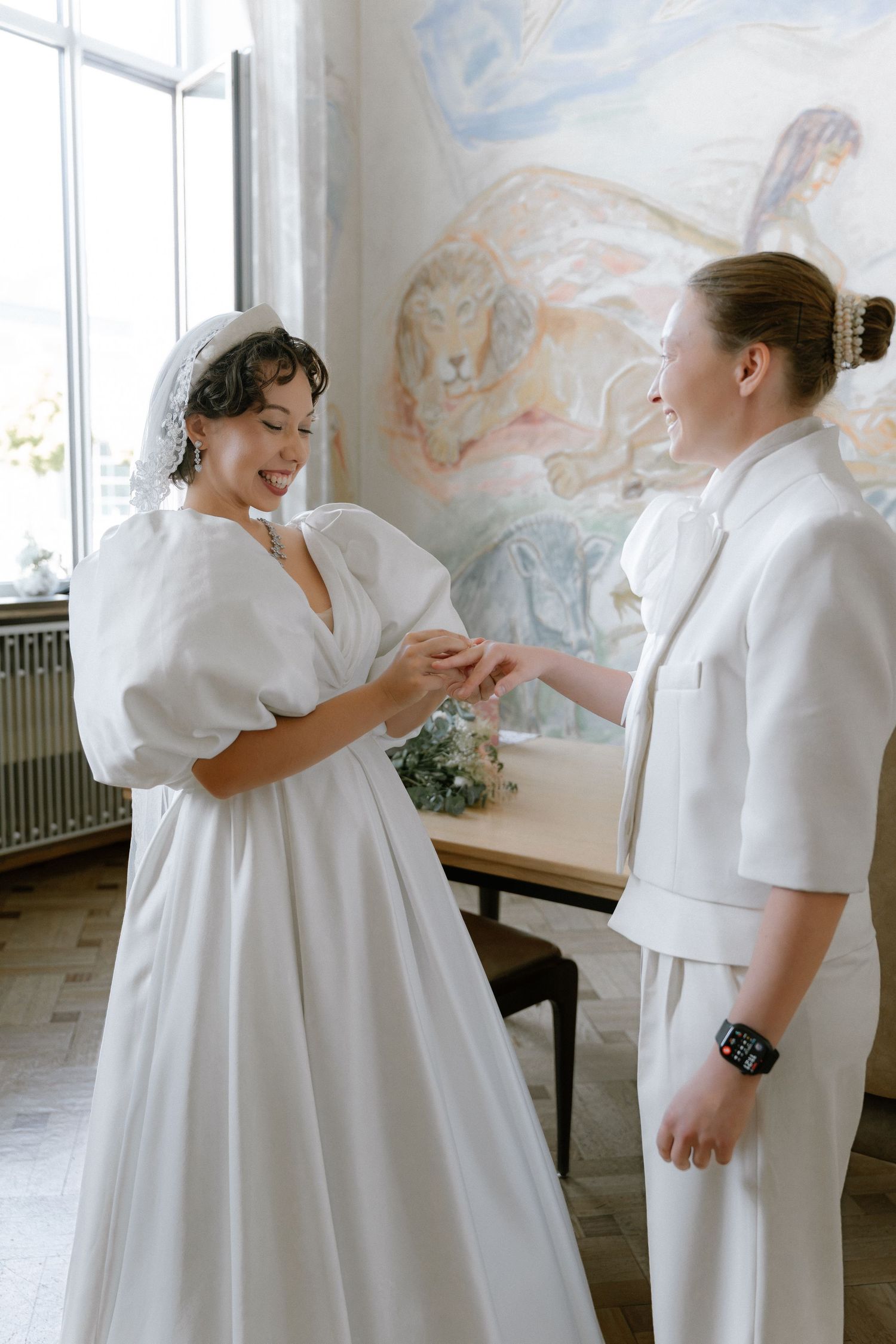 Two brides exchange rings in elegant white outfits during an intimate wedding ceremony indoors.